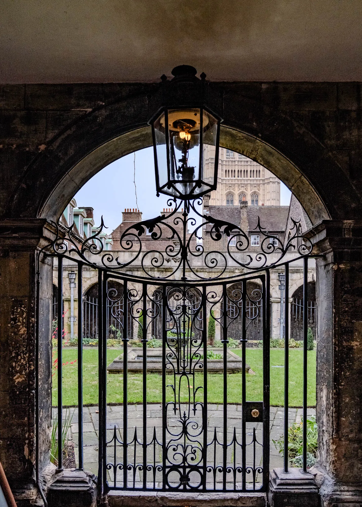 The image shows a view through an arched doorway. Above the doorway is a black, ornate metal lamp. The doorway leads to a small garden with a stone fountain and a stone walkway. The gate is made of black wrought iron. It has a intricate, ornate design. Behind the gate, a tall building is visible. It appears to be a church or a cathedral. The building has many windows and a tall tower with a spire. The stone walls of the doorway and surrounding buildings are old and weathered.