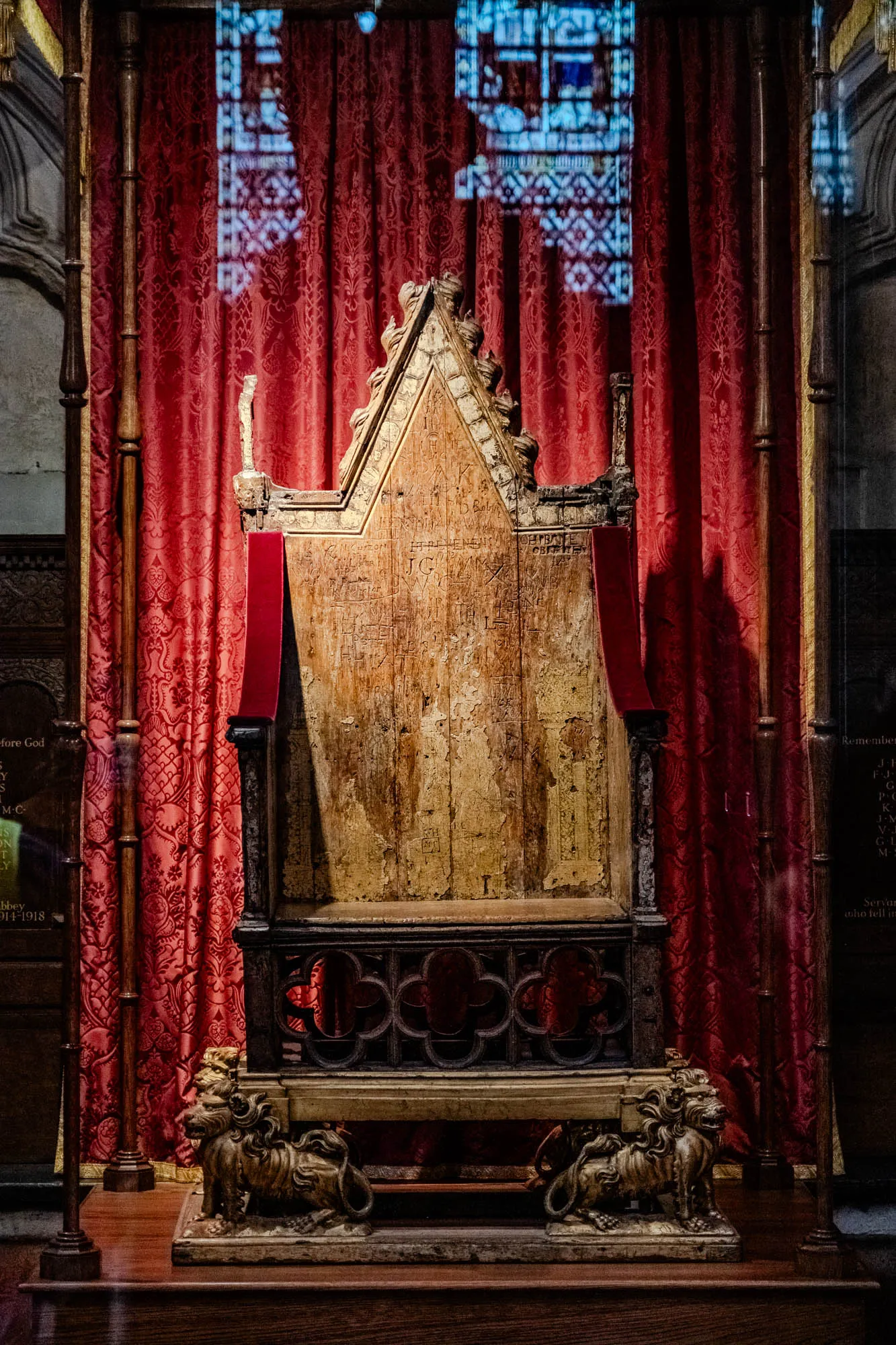 The image is of an ornately carved wooden throne, with an elaborate, tall, pointy back. The throne is set against a backdrop of red velvet curtains, with two golden lion statues on either side of the base. The throne is very old and worn, with many carvings and faded writing on the back and sides. The throne is lit from the front, creating a warm, inviting glow.