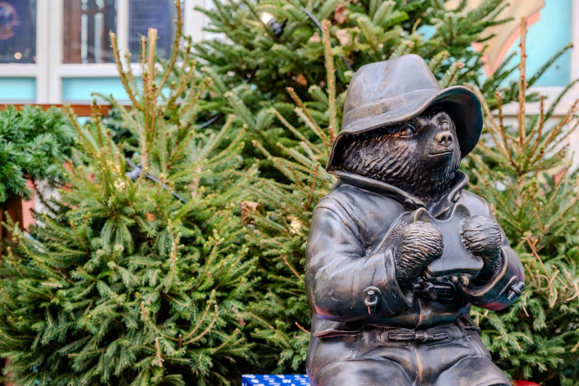 A bronze statue of Paddington Bear, standing in front of a Christmas tree. He is wearing his signature blue duffel coat and a brown hat. He has a  slightly serious expression on his face and his arms are crossed, holding what appears to be a small brown suitcase. The background is blurry but you can see a blue sky and a brick wall.