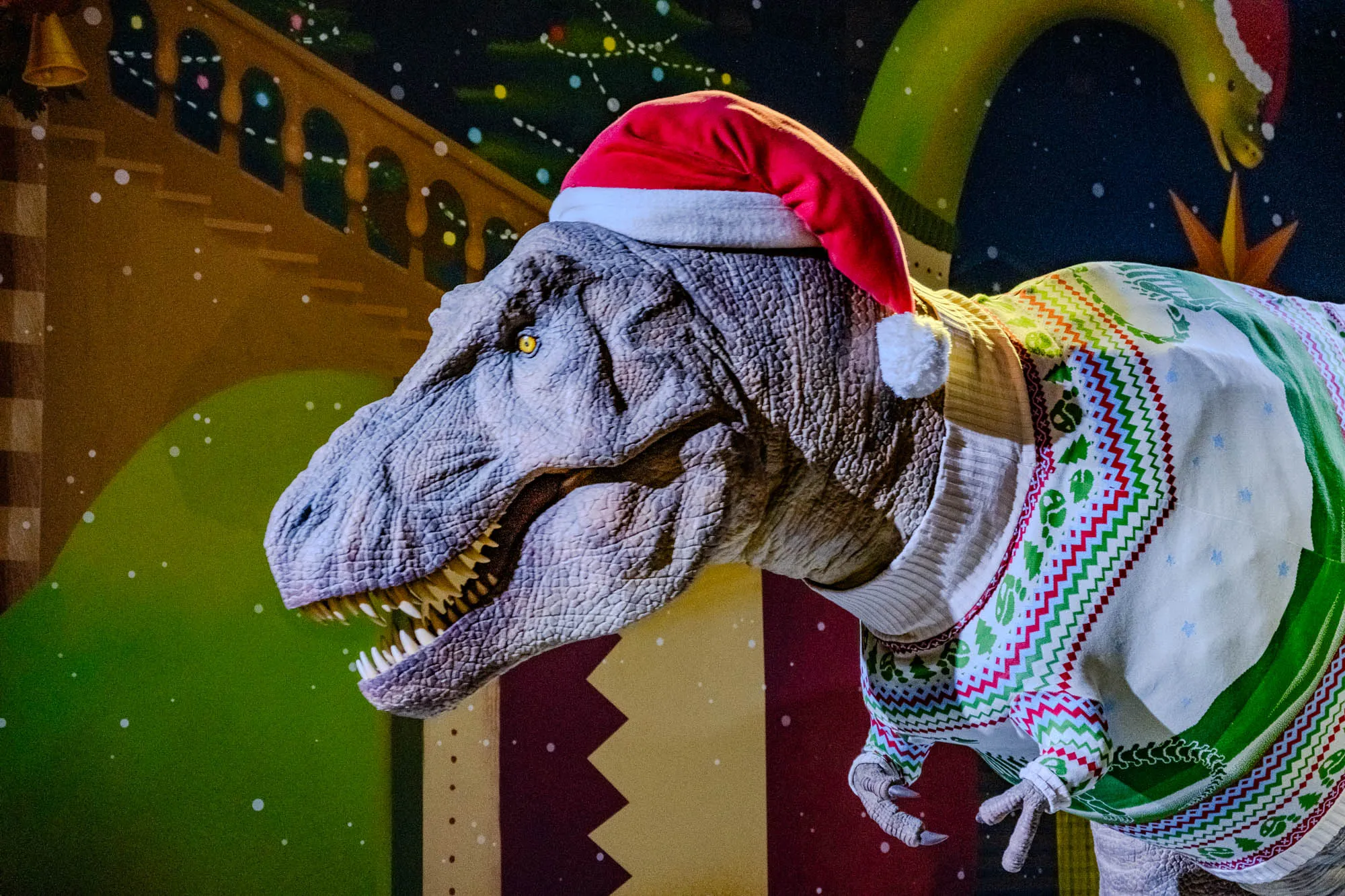 This image shows a T-Rex dinosaur wearing a red Santa hat and a white, green, and red sweater. The dinosaur's head is turned to the left and it's looking towards the viewer. The dinosaur is in focus and the background is blurred. The background is a colorful mural with green and brown tones and includes a Christmas tree with white lights and a green long-necked dinosaur in a Santa hat as well.  There are white dots in the background that represent snow.  The dinosaur's mouth is open and its teeth are visible.