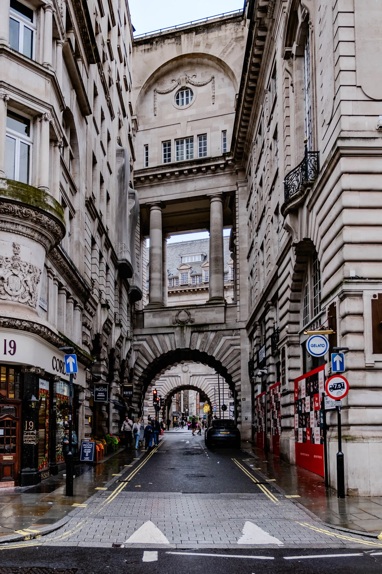 The image is a view down a narrow street in London. The street is lined with tall buildings, all with a similar architectural style. The buildings are made of stone and have many windows. The street is paved with cobblestones and there are yellow lines painted on the road to indicate the lanes. The buildings are all very old and there are some details on the walls, like decorations around the windows. There is a large arched structure at the far end of the street. The street is empty except for a few people walking and a car driving down the street. It is a gray day. There are a few street signs visible, with a blue "GELATO" sign visible on the right, and a sign indicating an arrow pointing straight up on the left. There is a sign for a shop called "Piggy's" on the left side of the image. There is also a white arrow painted on the road in the foreground.