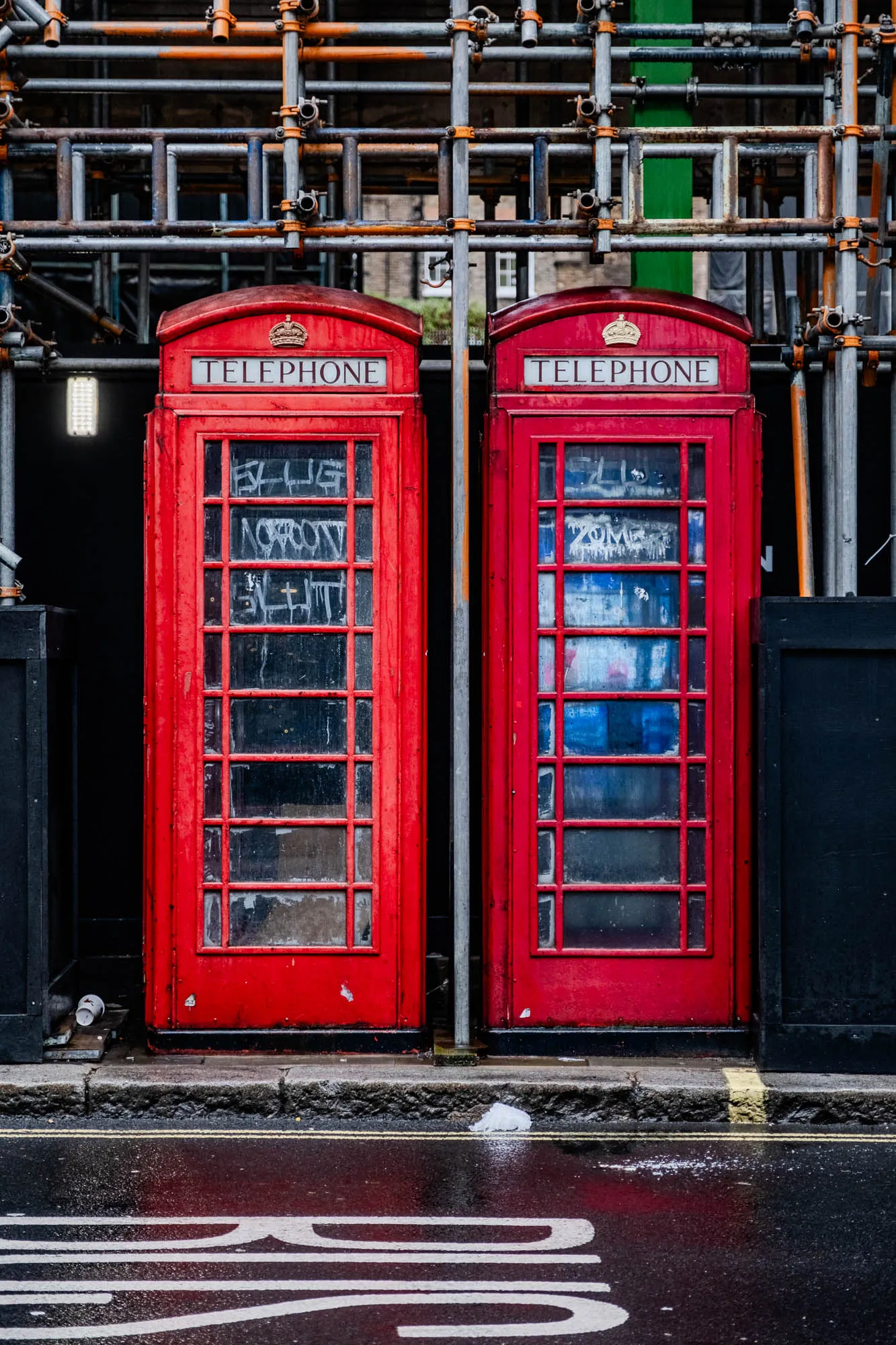 The image shows two classic red telephone booths, side-by-side, on a wet city street. They are both closed, but the glass panels of the doors are visible. There is a silver metal scaffolding structure behind the booths, and it appears to be under construction. The street is wet from rain, and a white object is lying on the pavement in the middle of the street. There is also a large white painted marking in the road that reads "BUS" in large letters.