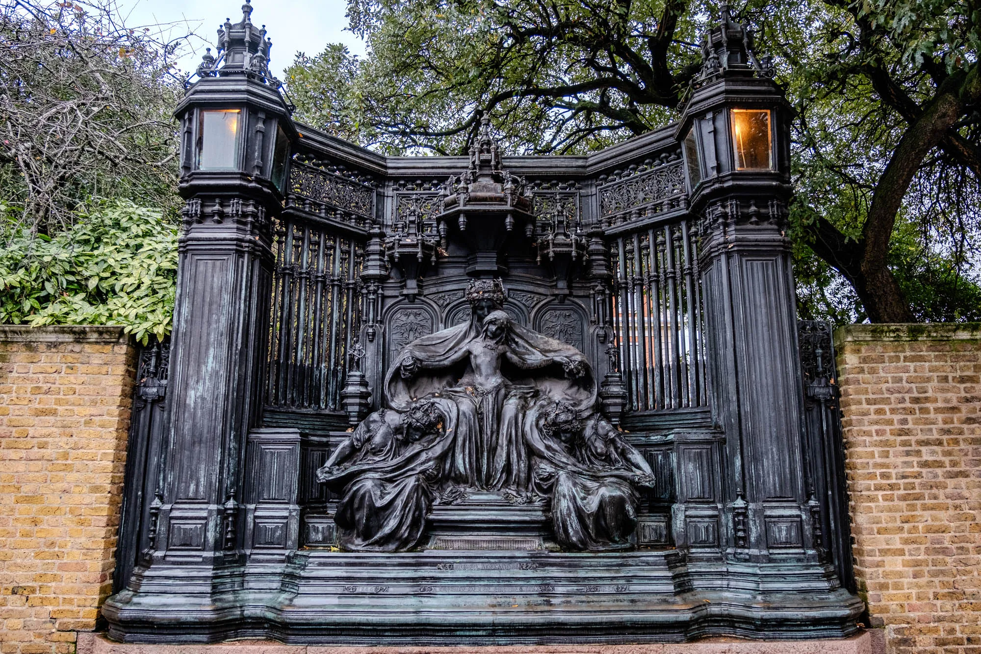 The image is of a large, intricate, black metal monument. It is made of black metal and features a tall, ornate gate with two large pillars on either side. The gate is decorated with a series of vertical bars and intricate carvings. There are two large lamps on the top of the pillars, one on each side, with a lit yellow light shining from each lamp. There is a large, elaborate metal sculpture that sits in the middle of the gate. The sculpture features a woman in flowing robes sitting on a throne. The woman is surrounded by two other figures. 

The sculpture is on a platform with a brick wall surrounding it. The brick wall is a muted yellow color and is made of brick. There are some small trees and branches in the background. The metal monument is in the foreground and has a weathered texture. 
