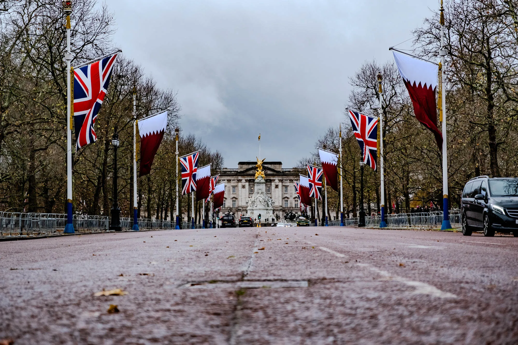 The image shows a street lined with trees and flags. There are several flags of the United Kingdom and a few flags of Qatar, all on tall white poles. The street is wet and looks to be cobblestone. In the distance is a large building with a statue of a golden figure on top. There are several cars on the road and a few people walking in the background. There are fences along the sides of the road with blue accents, and a few small trees and plants can be seen in the background. The sky is overcast and appears to be gray. The image was likely taken on a day after a rain shower.  The image focuses on the road leading towards the building, giving a sense of perspective.  The image is taken at a low angle, which emphasizes the height of the flags and the size of the building in the distance.  The flags are slightly blurred in the distance, giving a sense of depth.