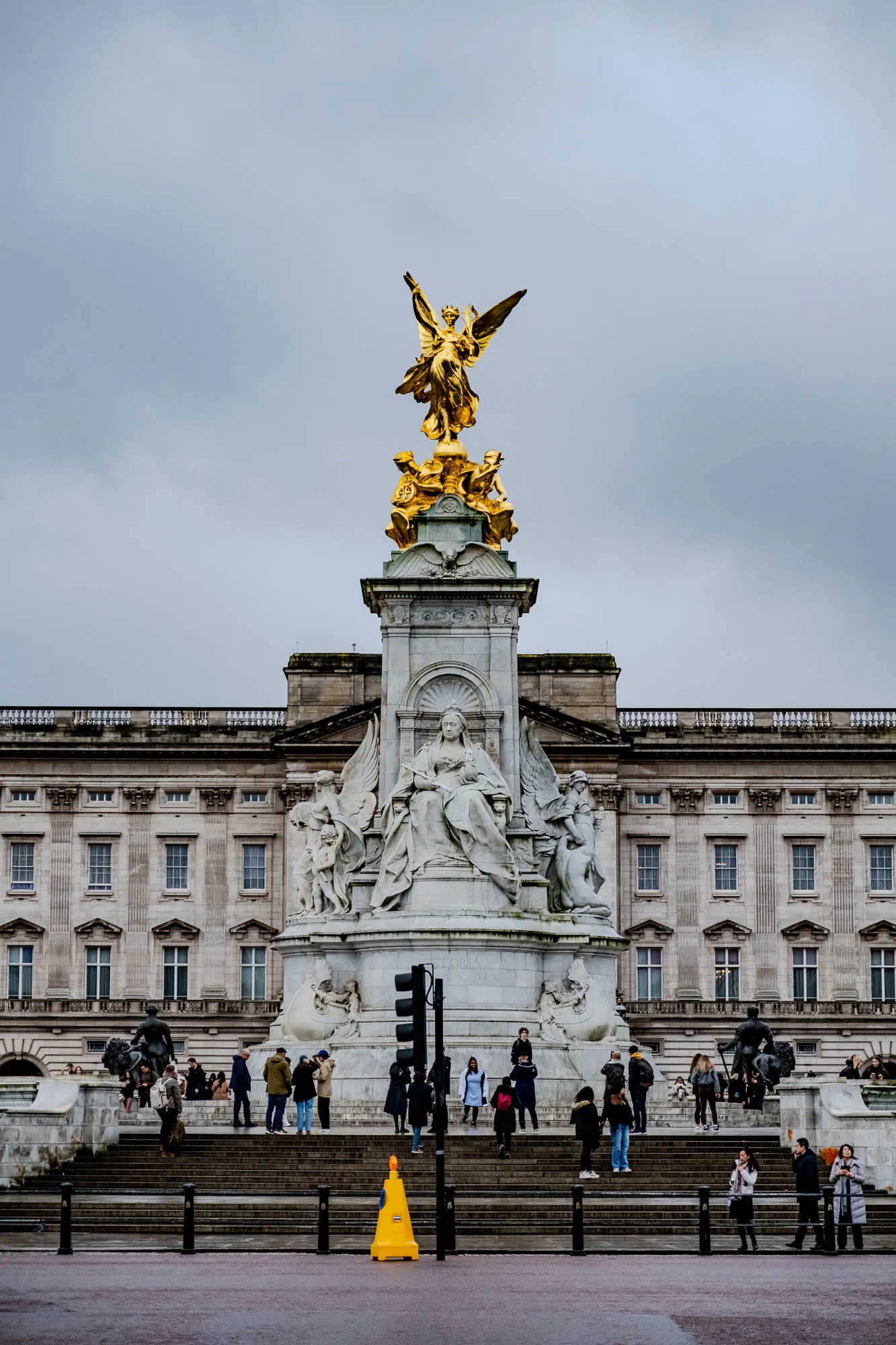 The image shows the Victoria Memorial in front of Buckingham Palace. The monument is a large, ornate structure with a golden statue of Queen Victoria at the top. The statue is surrounded by four bronze groups representing different aspects of Victoria's reign. There are many people in the foreground, walking up the stairs to the memorial. The sky is overcast and the ground is wet, suggesting it may have been raining recently.  There is a yellow construction cone in the foreground, along with a traffic light that appears to be on a timer.  Buckingham Palace is a large, white building with many windows, but there are no details or features that can be described in detail. It is possible that the photographer was positioned at a relatively low angle and only the front facade of the palace is visible.  The image captures the grandeur of the Victoria Memorial and the beauty of Buckingham Palace in a dignified and somber way.  The people in the foreground add a human element to the image, while the overcast sky creates a mood of quiet contemplation.  The yellow construction cone acts as a point of interest and creates a sense of scale. 
