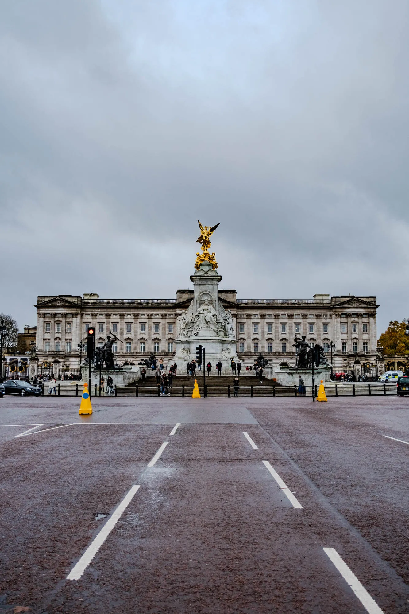 The image shows a view of Buckingham Palace from the road in front of it. In the center of the image is a large monument with a golden statue of a woman holding a crown and a scepter on top. The monument is surrounded by a large circle of sculptures. The palace is a large, rectangular building with many windows. There are people walking in front of the palace and monument. There are yellow traffic cones on the road in the foreground. The sky is cloudy. The pavement appears wet.