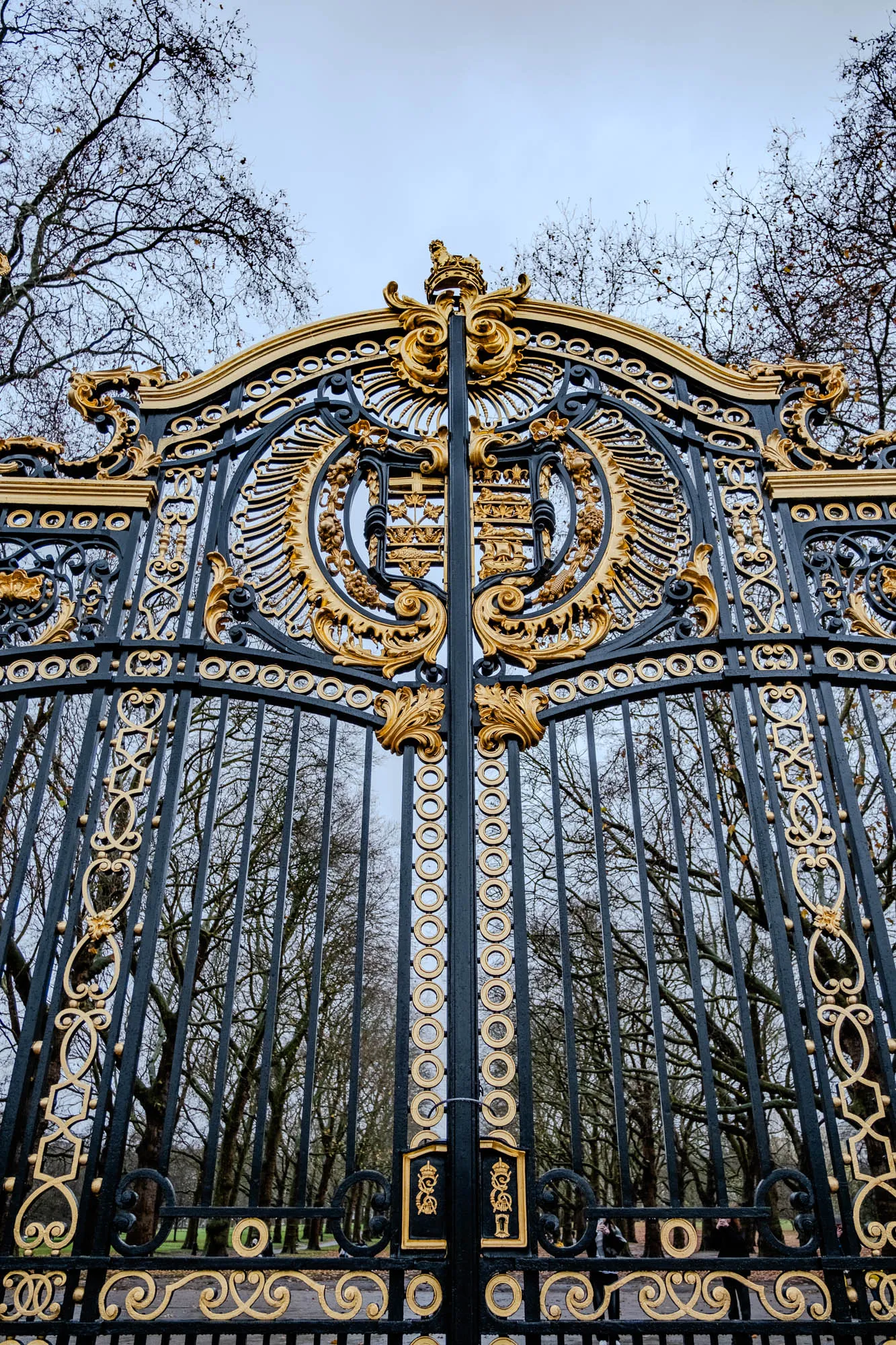 This image is a close-up of a large, ornate gate. The gate is made of black metal, with gold accents. It is made of vertical bars with elaborate goldwork and rings around the bars. The gate has two large panels that meet in the middle.  The top of the gate is a decorative crest with a gold crown. The gate is set against a backdrop of bare trees and a cloudy sky. The gate is open, and you can see a path leading through a park behind it.  There are two people walking through the gate.