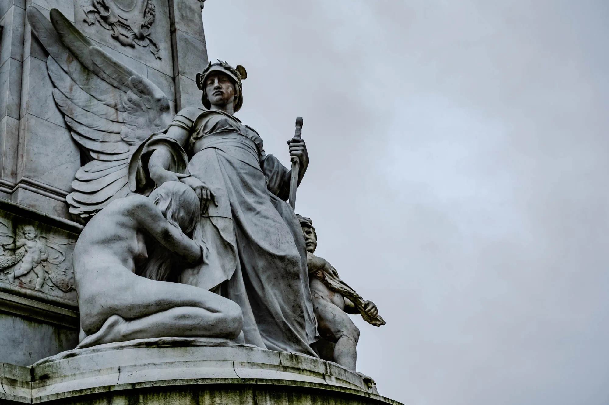 The image depicts a stone statue of a winged woman with a sword in her right hand. She is wearing a flowing gown and has a crown of leaves on her head. She is standing over a smaller figure kneeling at her feet, with her head bowed and her arms wrapped around herself. There is a third figure to the right of the central figure, appearing to be male, and also holding a weapon. The statue is set against a plain, light grey cloudy sky. The statue is made of rough-textured stone, possibly marble, with visible signs of weathering. The statue appears to be part of a larger monument.