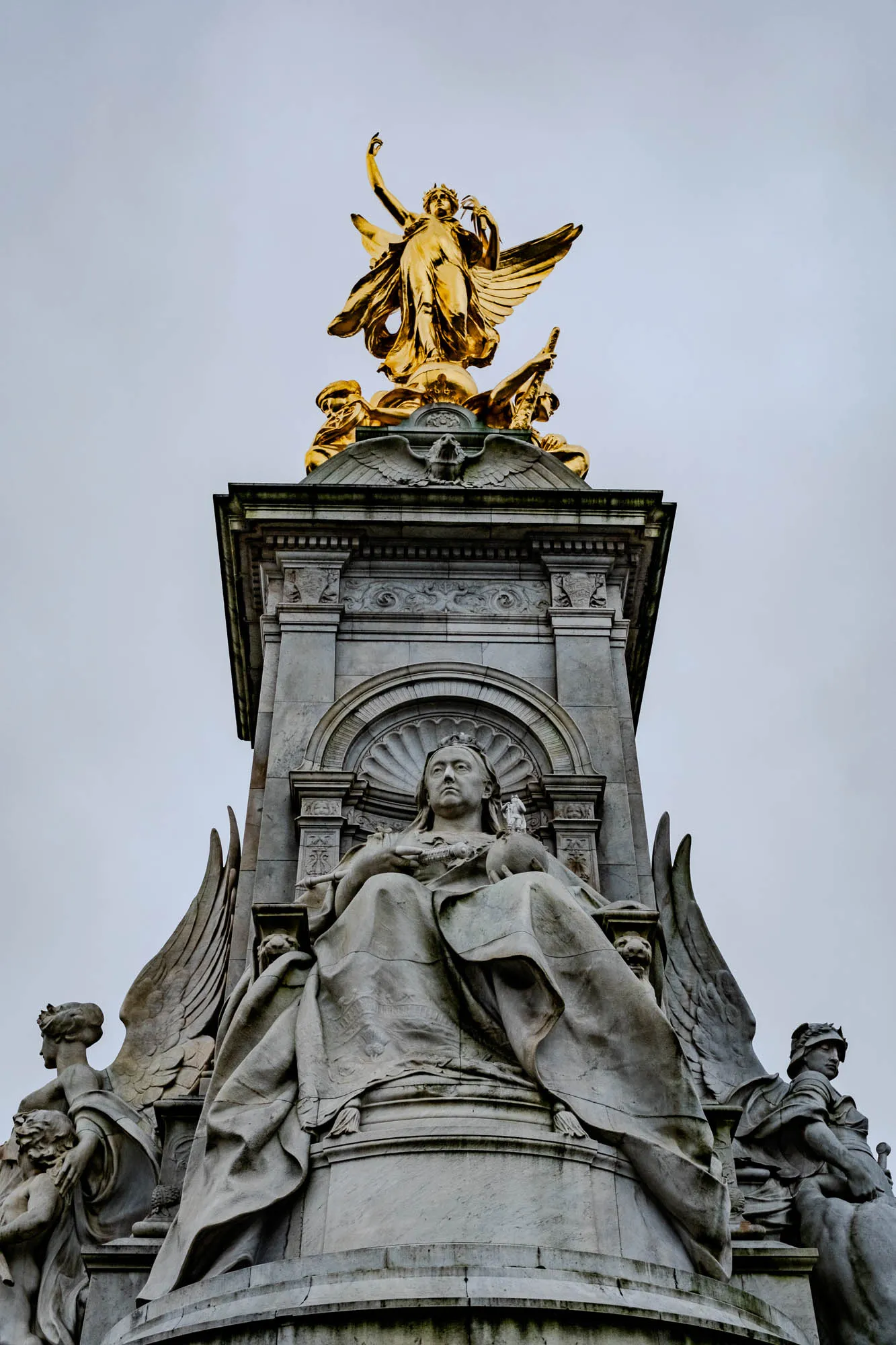 This image shows a large stone monument, a memorial to Queen Victoria.  The Queen's statue is seated at the center of the monument, wearing a crown and robe, with her hands resting on her lap.  The statue appears to be looking to her right.  She sits on a stone throne with a large, sculpted angel's wing to the right of her.  The wing is attached to the throne.  To her left are the outlines of another wing that is partially hidden from view.  Above the statue is an ornate arch, and at the very top of the monument is a golden statue of a woman with wings.  The woman is standing on a large globe.  She is holding a small wreath or bouquet of flowers in one hand.  Below her, there are two smaller statues, and the entire top of the monument is ornate.  The sky in the background is gray and cloudy.