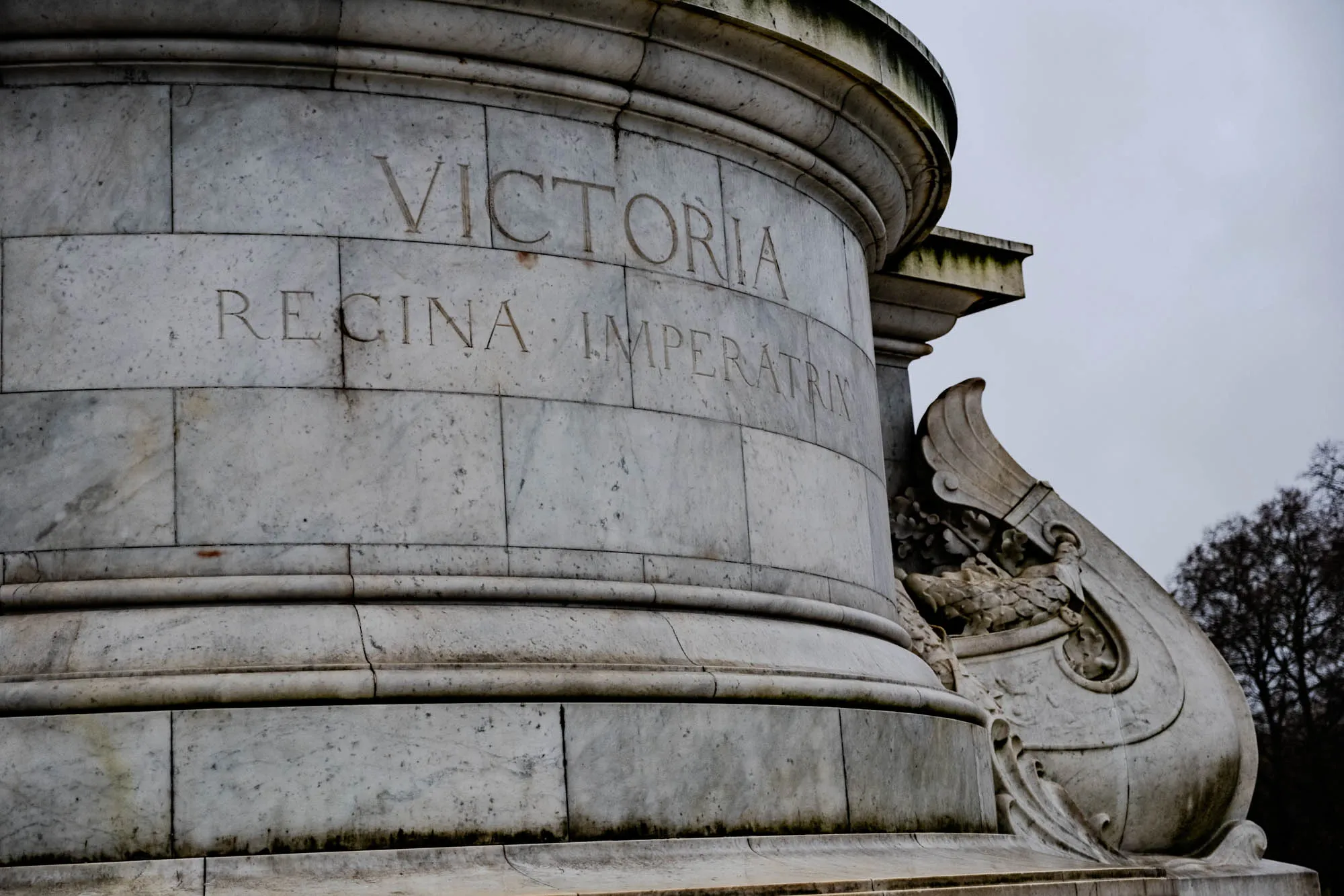 This is a close up of the base of a marble statue, with the inscription "Victoria Regina Imperatrix".  The base is made of large blocks of white marble, and is intricately carved with details.  The inscription is worn, but still readable, and the base is weathered with moss.  The base of the statue is decorated with a decorative, carved design that flows around the side and blends with the main structure. The statue is against a cloudy, gray sky, and some trees are visible in the background.  The statue appears to be old. 
