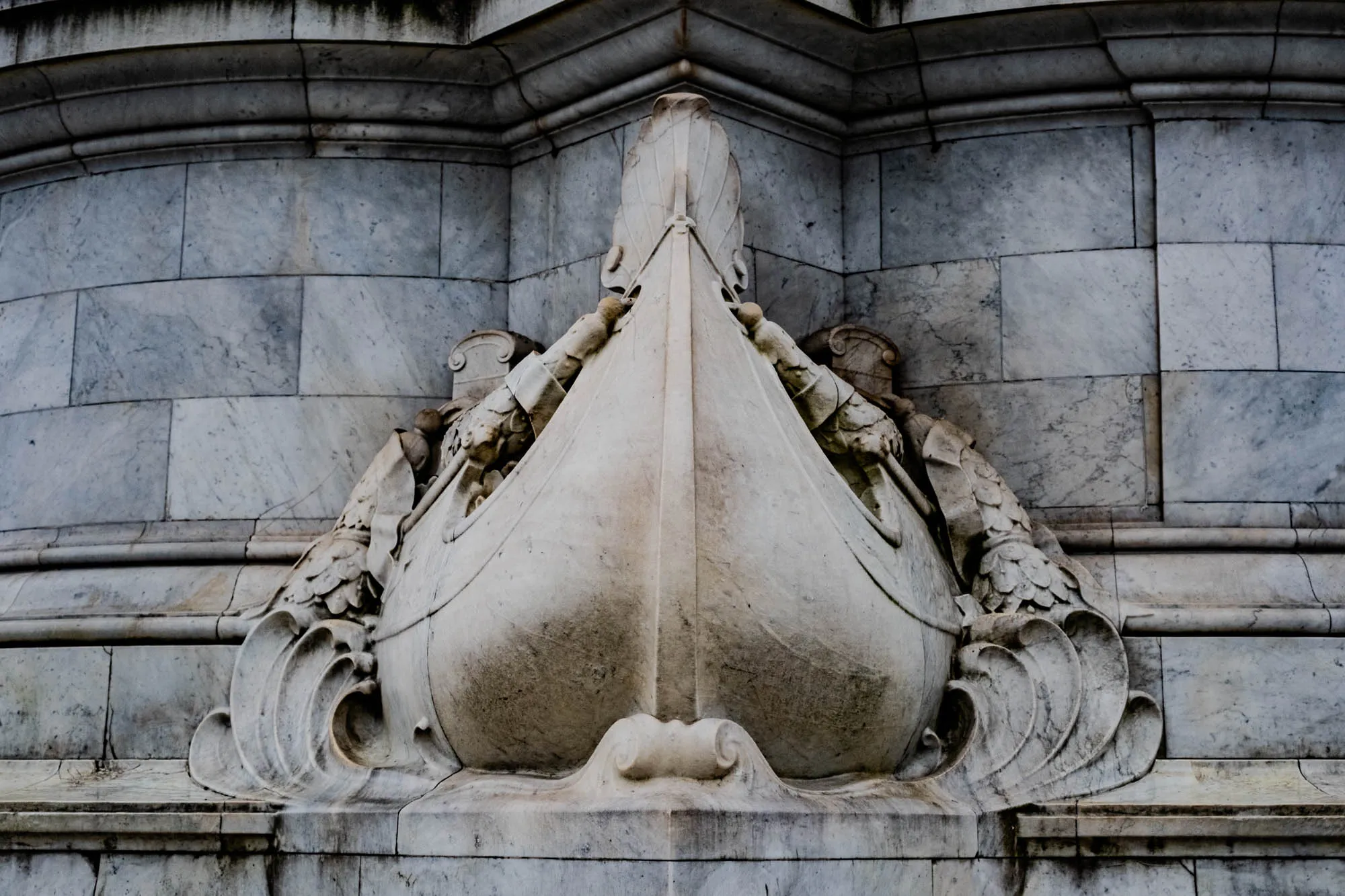 A close-up of a stone sculpture of a boat with a row of stone blocks above and below the boat. The boat appears to be carved out of the stone block and has a rounded bow. There are decorative flourishes on either side of the boat in the stone representing the boat moving through the water. The background is a rough texture and the stone is a mottled grey.