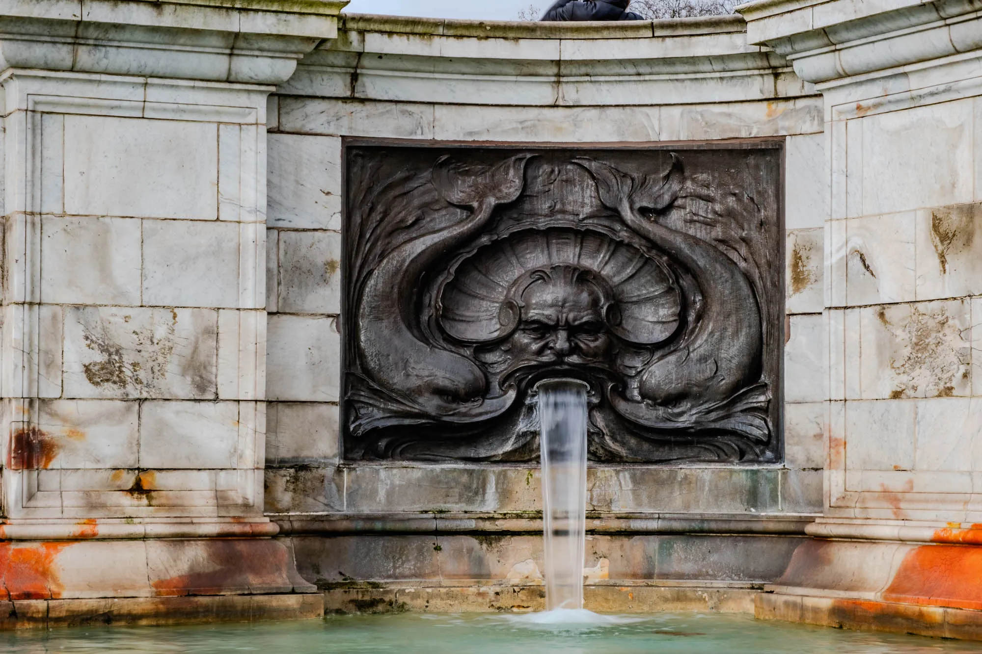 This image shows a fountain made of stone. The fountain has a carved stone face that is part of a larger, elaborate design. Water flows from the mouth of the carved face, and the water cascades into a pool of water in front of the fountain. The fountain is made of a white stone, and the stone is worn and has patches of orange and brown. There are two large stones on each side of the fountain.