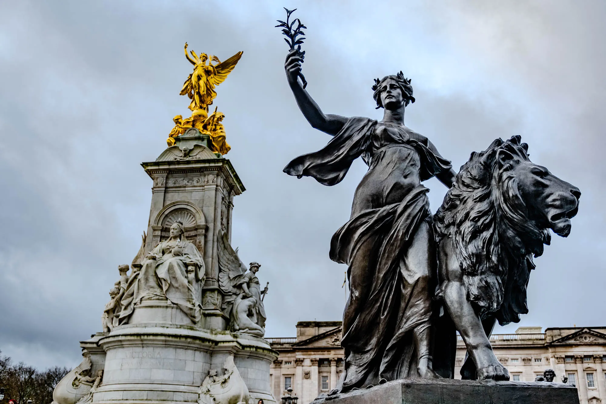 The image shows a statue of a woman riding a lion, the woman is holding a branch of leaves in her right hand, the statue is in the foreground with a large white monument in the background, the monument has a statue of a woman with wings on top, the woman in the background is holding a sword, the background also has a large white building with many windows.  There are trees to the left of the image. The sky is cloudy. The statue of the woman riding the lion is made of bronze. The statue of the woman with wings is made of gold. The monument is made of white stone. The building in the background is made of white stone.