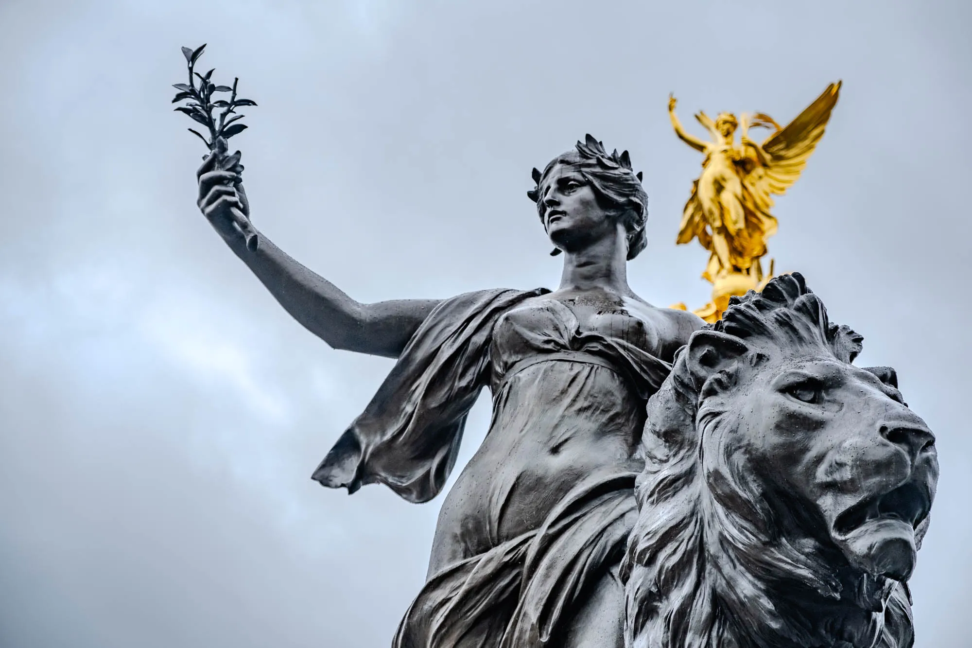 The image is a close-up of a statue of a woman, likely representing Peace or Victory, standing on a lion. She is holding a branch of an olive tree in her right hand, a symbol of peace. Her left hand is outstretched. She is wearing a flowing robe and a crown of leaves. She has a calm expression on her face. In the background, another statue of a woman in a flowing robe is partially visible, made of golden metal. She is standing on a raised platform above the first statue, looking down at the viewer. The sky is gray and cloudy.  The statues are likely part of a monument or memorial.