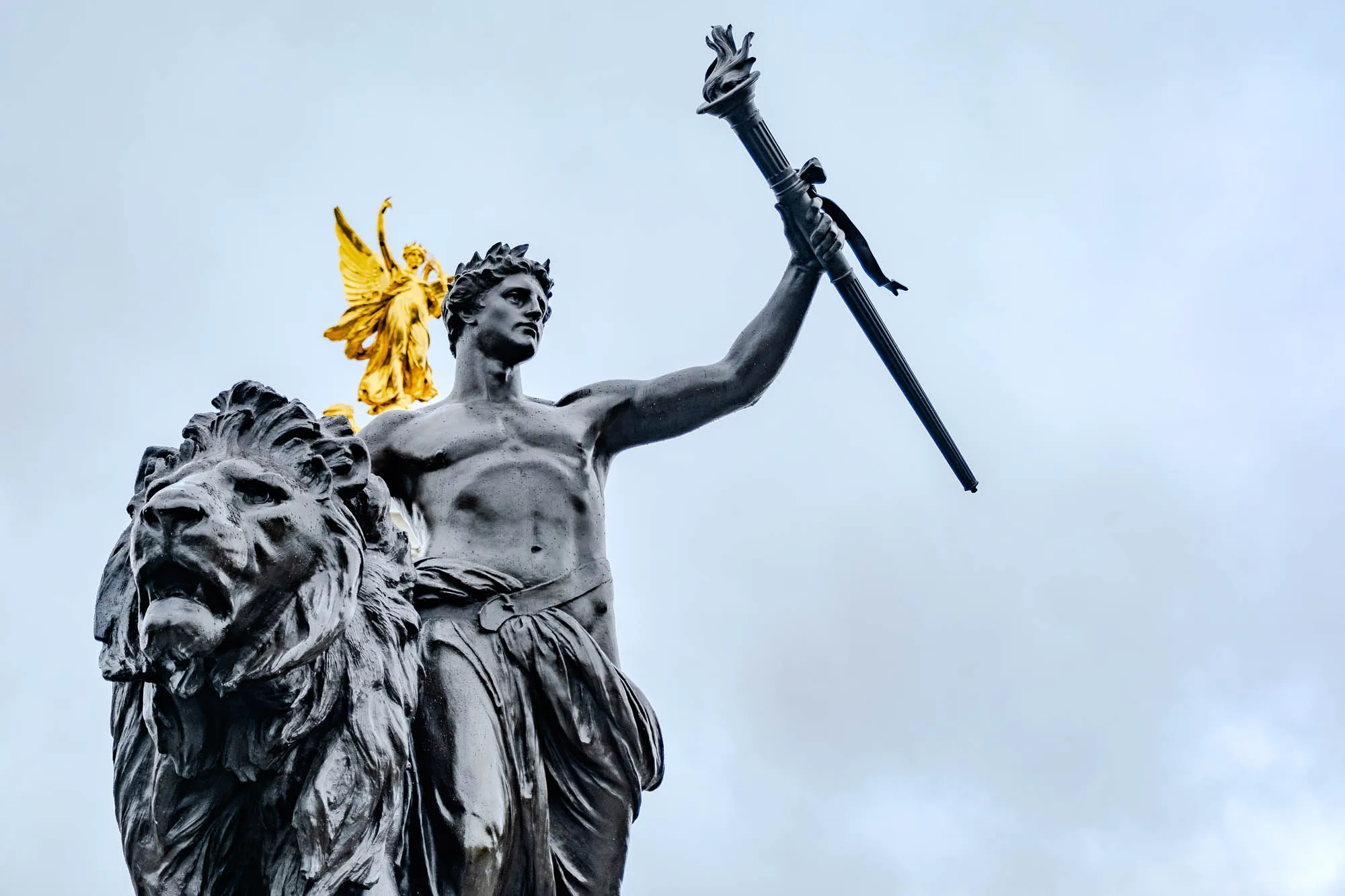 The image shows a black and gold statue of a man, possibly a soldier, holding a torch in his right hand. The man is standing on top of a lion and has a golden angel behind him with wings spread out. The statue is in front of a light blue cloudy sky. The statue is made of metal, probably bronze, and the torch and angel are made of gold. The man is wearing a long robe, possibly a toga, and is holding the torch with his right hand, while his left hand is on the lion's back.  The lion is looking towards the left, while the man is looking towards the right.