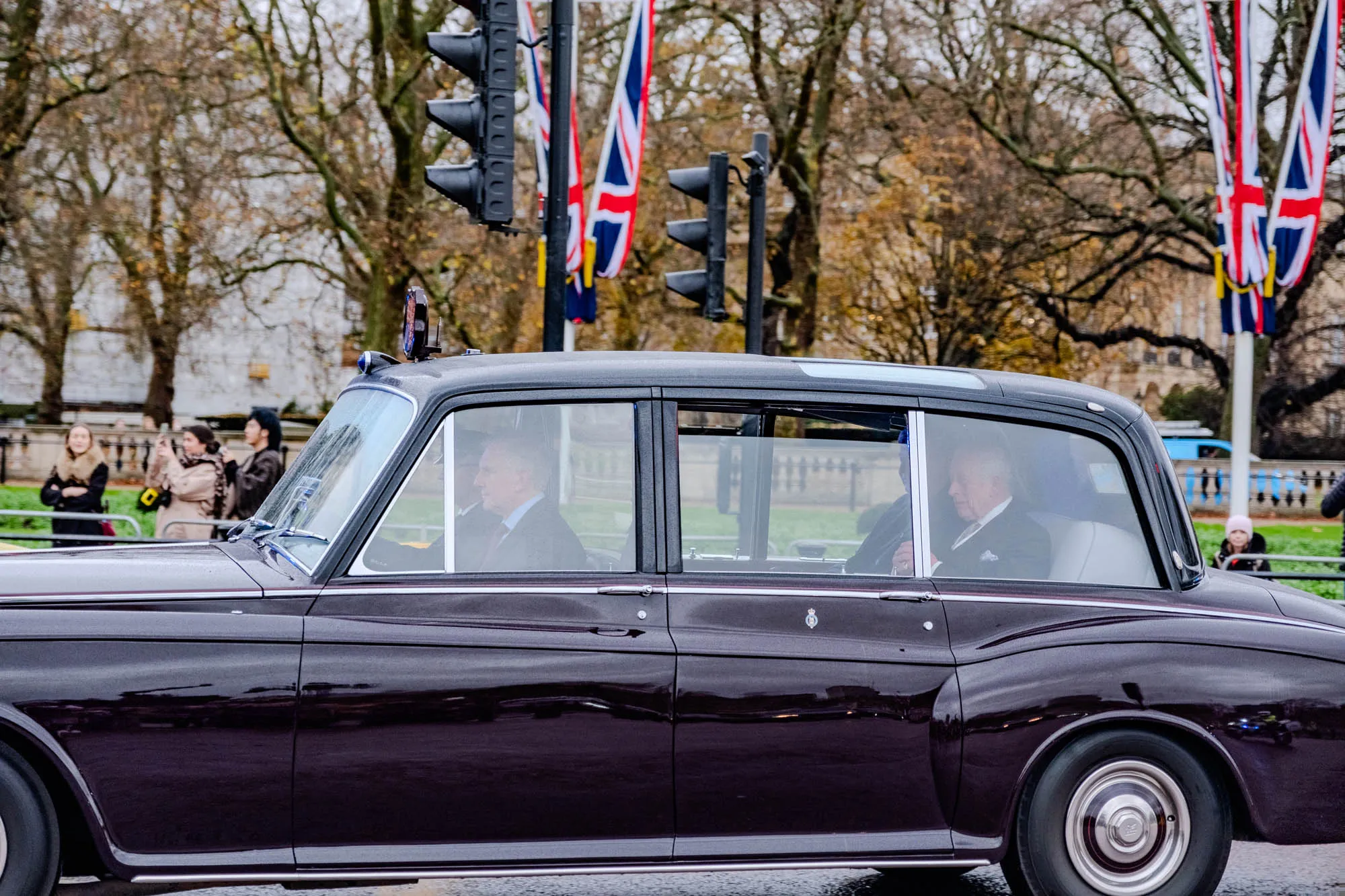 The image shows London black cab. The car is a sedan with a long, sleek hood and a spacious interior. The windows are tinted, and there are two people sitting in the back. The car has a shiny, polished finish and large chrome hubcaps. There is a small crown and symbol on the passenger door.

The two people in the back are older white men, both wearing dark suits and light-colored shirts.  The man on the left is facing forward and has his hand on the door. The man on the right is also facing forward and has his hands in his lap. It is possible they are both wearing ties. They are not clearly visible because of the tinted windows.

The car is being driven on a street with a gray surface.  On the right side of the image, there are three British Union Jack flags on poles.  There are traffic lights in the background, and the car is stopped at a traffic light. In the background, there are trees, likely in an urban environment.  There are also a group of people, likely pedestrians, standing to the left of the car. The people are unclear, but they are all likely wearing dark clothes. They are blurred and appear to be looking at the car. They may be taking photos of the vehicle.