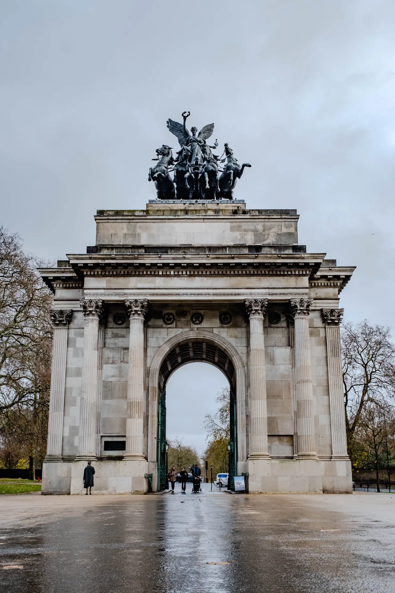 The image shows a large stone archway with a statue on top. The statue depicts a woman in a chariot pulled by four horses. The archway is made of white stone with a large arch in the middle. The archway is surrounded by four large columns on each side. The image is taken from a low angle looking up at the archway. The sky is overcast and there are trees to the left and right of the archway. The ground is wet and reflective. A group of people are walking through the archway.