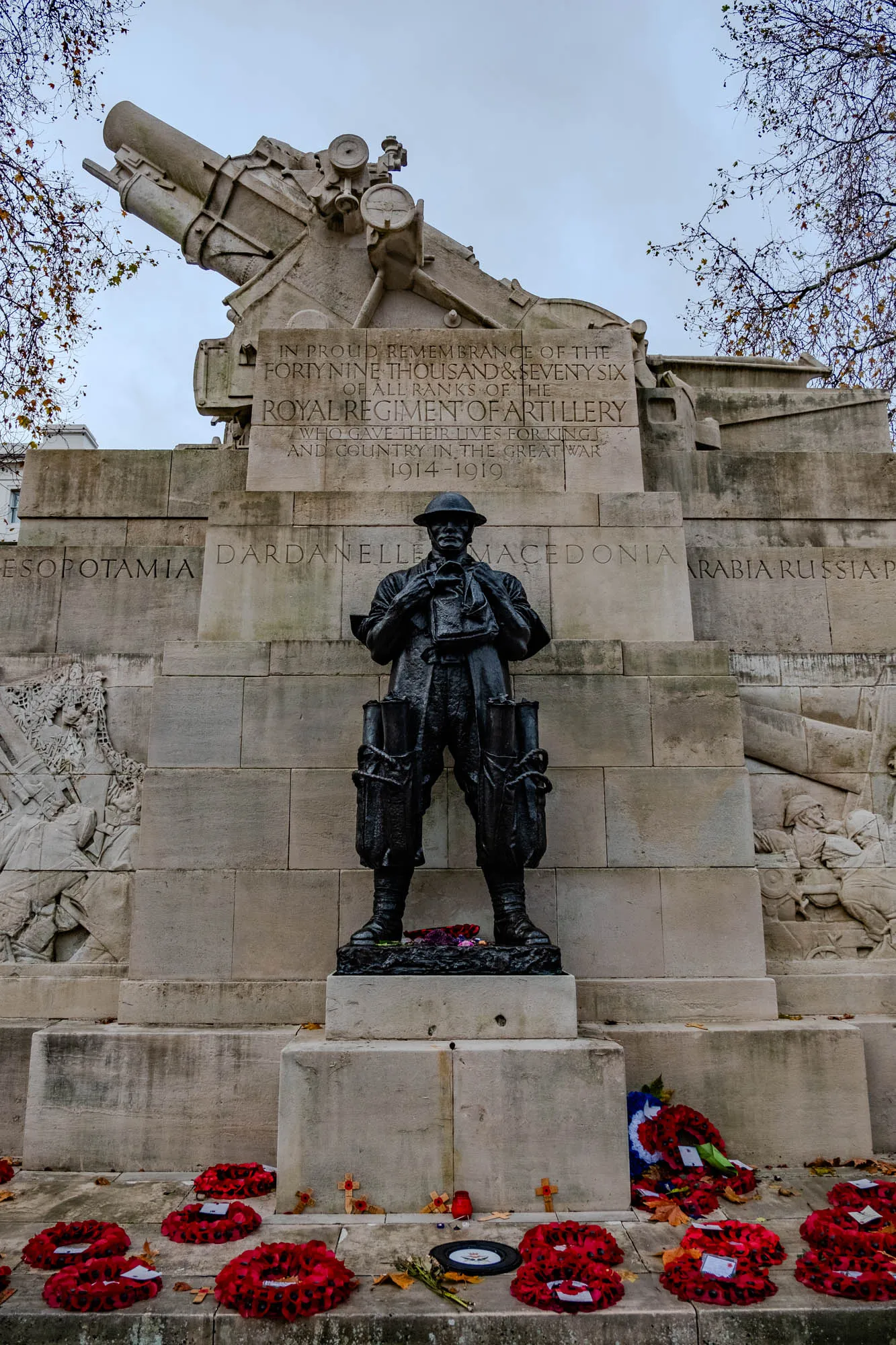 A stone monument with a cannon on top and a statue of a soldier in front. The soldier is wearing a uniform and a helmet, and he is holding a rifle. The monument is inscribed with the words "IN PROUD REMEMBRANCE OF THE FORTY NINE THOUSAND & SEVENY SIX OF ALL RANKS OF THE ROYAL REGIMENT OF ARTI LLERY WHO GAVE THEIR LIVES FOR KING AND COUNTRY IN THE GREAT WAR 1914-1919". The monument is surrounded by wreaths of red poppies. The monument commemorates the soldiers of the Royal Regiment of Artillery who died in World War I. The words "ESOPOTAMIA DARDANELLE MACEDONIA ARABIA RUSSIA-P" are inscribed on the side of the monument.