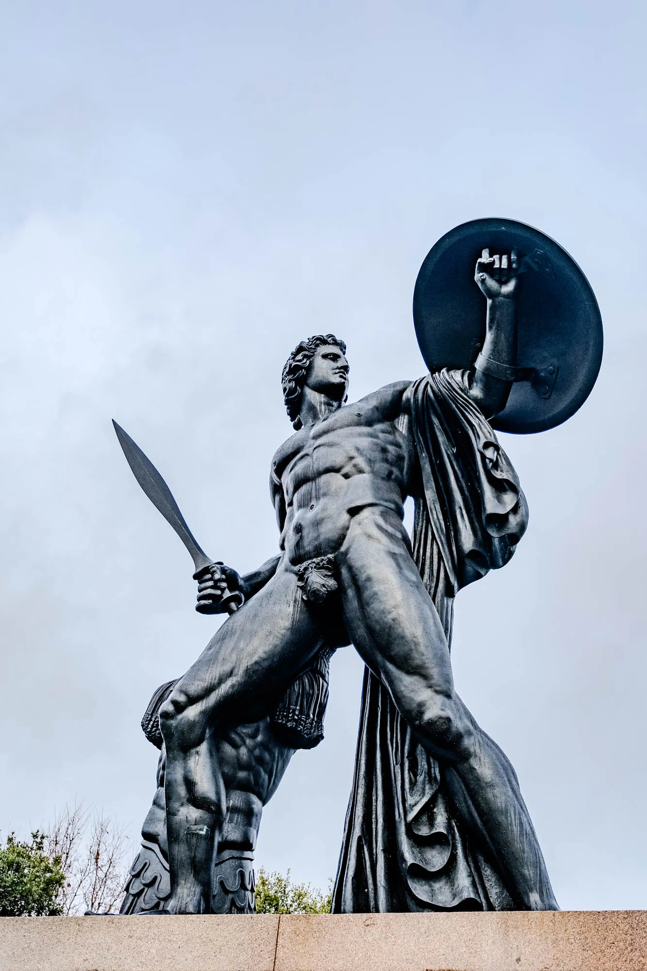 The image shows a bronze statue of a muscular man holding a shield in his right hand, and a sword in his left hand. He is wearing a long, flowing robe and is standing on a stone pedestal. The statue is set against a cloudy sky. The man is facing to the right of the viewer, and his right arm is raised above his head, holding the shield. The shield is round and has a raised edge.  The man's left hand is holding a sword, which is pointing down at his left side.  The man's robe is flowing down his back and around his legs, covering most of his body.  The statue is very detailed, with realistic musculature and facial features. The background is a simple, cloudy sky. 
