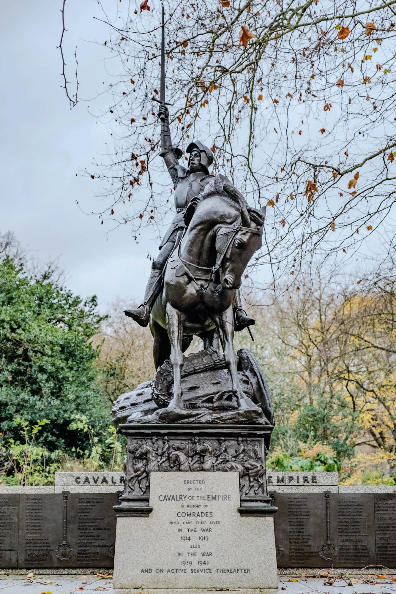 The image shows a bronze statue of a knight on horseback, set against a backdrop of a park with autumnal trees in the background. The knight is standing on a platform, holding a sword aloft, with the horse's head turned towards the viewer. There is a plaque on the platform in front of the statue, which reads: "Erected by the Cavalry of the Empire, in memory of comrades who gave their lives in the war 1914 1919, also in the war 1939 1945 and on active service thereafter."  There are three other plaques on either side of the statue’s platform, with the words "Cavalry" on the left and "Empire" on the right. The plaques are filled with the names of individuals. The statue and plaques appear to be a war memorial.  The image was taken on a cloudy day and the trees are mostly bare, with some brown leaves on the ground.  The image is taken from a slightly low angle, looking up at the statue. 
