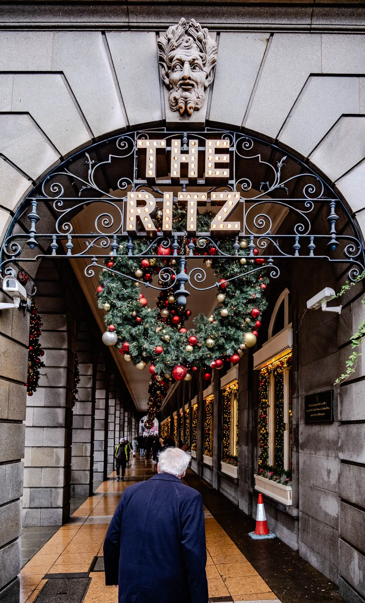 The image shows a sign that says "The Ritz" in large illuminated letters, with a stone sculpture of a man's head with a beard and a wreath of evergreen branches decorated with red and gold ornaments hanging from the sign. The sign is above a walkway with columns on either side, and the windows are decorated with Christmas lights. A man in a dark coat is walking down the walkway. There is a red and white traffic cone on the right side of the walkway. The walkway is paved with a mix of stone and lighter brown tiles, which appear wet and slightly muddy. There is a small green plant growing on the right side of the wall.