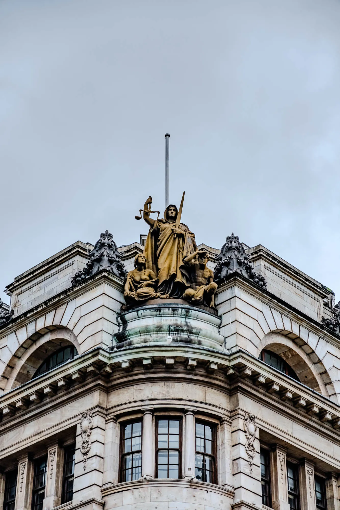 The image shows a close up of a building's corner, with intricate stonework. There is a statue of a woman holding a scale and a sword at the top. The woman's face is obscured by the hood of her robe, but she is holding a sword in her right hand and the scale in her left. To the woman's left is a statue of a woman holding a shield with her left hand. To the woman's right is a statue of a man holding a shield. The statues are facing forward. The building is made of stone and has an arched window on each side of the corner. The building is decorated with ornate carvings. The sky above the building is cloudy and overcast.