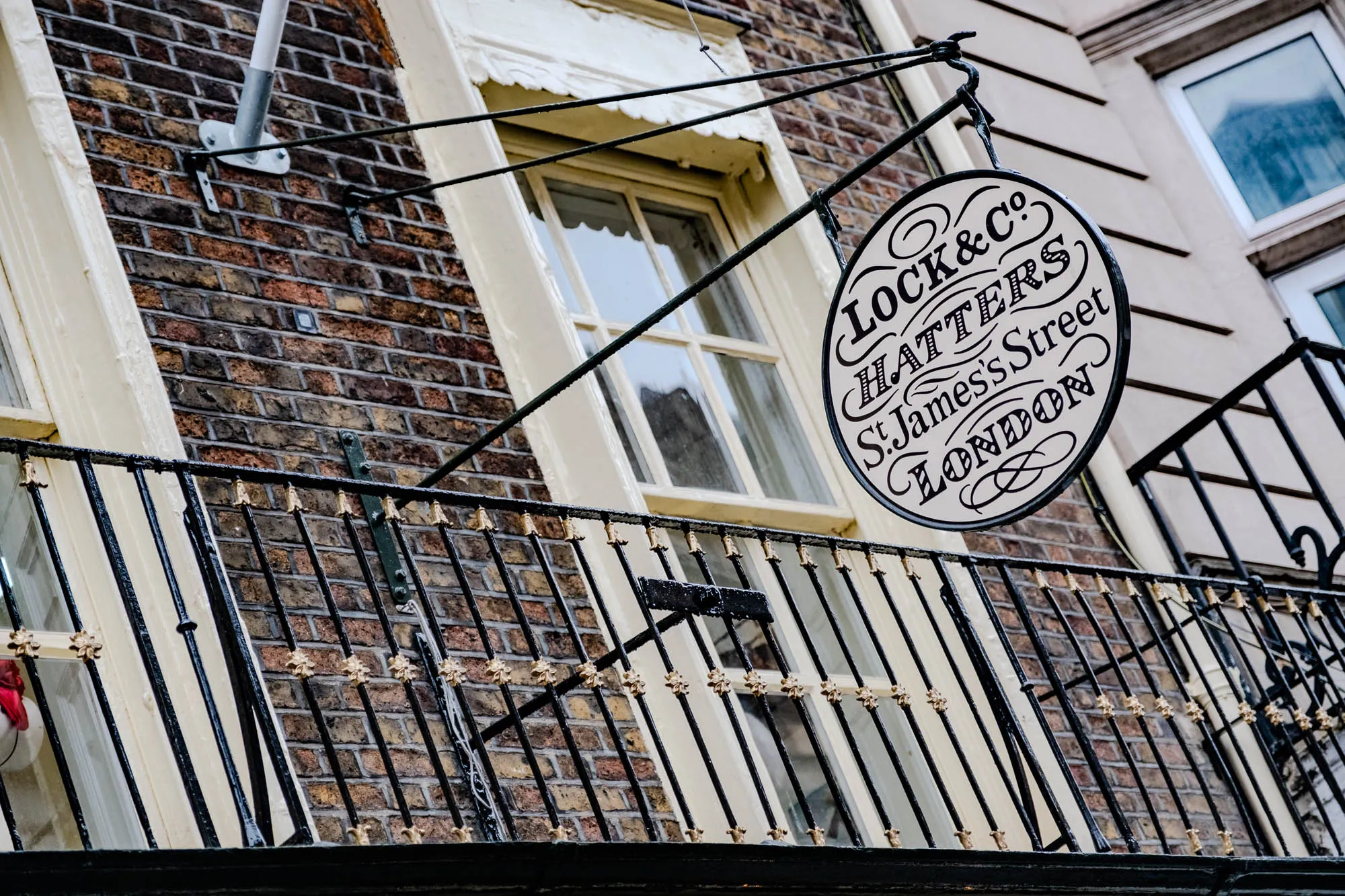 A close-up of an ornate, circular, white sign hanging from a wrought iron balcony. The sign reads, "Lock & Co., Hatters, St. James's Street, London" in a script font. The sign is hanging from the top of the balcony, which is attached to the front of a brick building. The building is old and has an aged facade.  The balcony has a black wrought iron railing with decorative, golden fleur-de-lis accents. The image is taken from a slightly low angle, looking up at the sign and balcony.