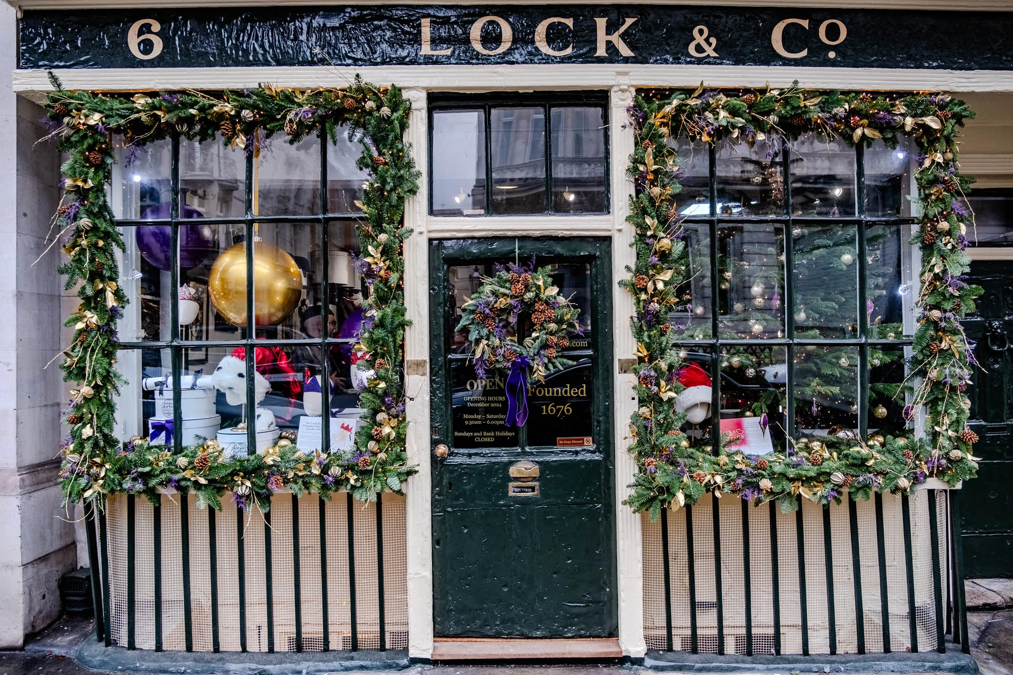 The image shows a storefront with a green door and two windows. The storefront is decorated with greenery, including pine boughs and garlands. The windows are decorated with garlands of greenery, pine cones, gold ornaments, and purple flowers. The greenery is interspersed with gold ribbon. There is a golden globe in the left window. The left window also has a white fuzzy item, possibly a stuffed animal. The door is dark green with a small sign that says “Founded 1676”. Above the door is a wreath of pine boughs, pine cones, and purple flowers. The door is closed.  The top of the storefront is black and has the number six and the text “LOCK & CO” in gold lettering. The bottom of the storefront has a black metal fence.  The storefront is in front of a white building.