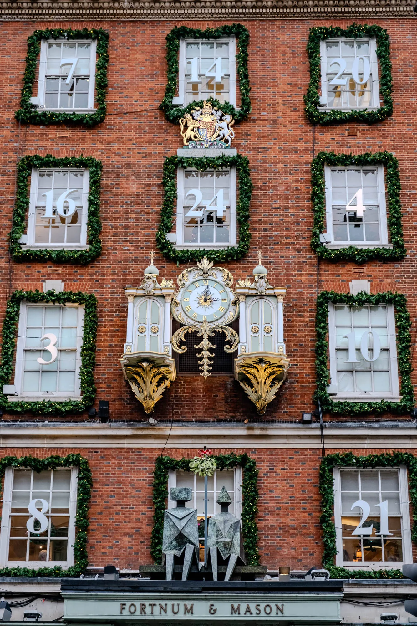 This is a photo of a brick building decorated for Christmas. The windows have white wreaths of greenery around them, and white numbers are placed in each window. The numbers visible are 7, 14, 20, 16, 24, 4, 3, 8, 10, and 21. Below the windows are two ornate clocks that appear to be gilded in gold with a white face. The clock on the left has a round face, and the clock on the right has a rectangular face. The building is adorned with a large wreath of greenery and a gold crest above the two clocks.  The building is topped with a cornice and a small sign that says "FORTNUM & MASON."  In front of the building is an abstract statue of two figures.