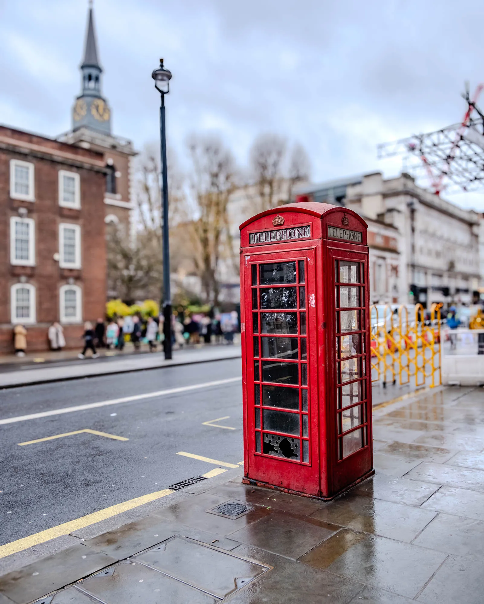 The image shows a red telephone booth on a city street. The booth is classic British, with a crown on top and the word "telephone" written on both sides. The booth has a glass door and windows, and it is sitting on the sidewalk in front of a tall building that's out of focus. The sidewalk is wet and there are yellow markings on the road. There's a street lamp behind the booth. The booth has a slightly faded paint job with a few small holes in the glass panels.  The street is empty except for some blurry people walking in the background. The sky is cloudy and the weather appears overcast.