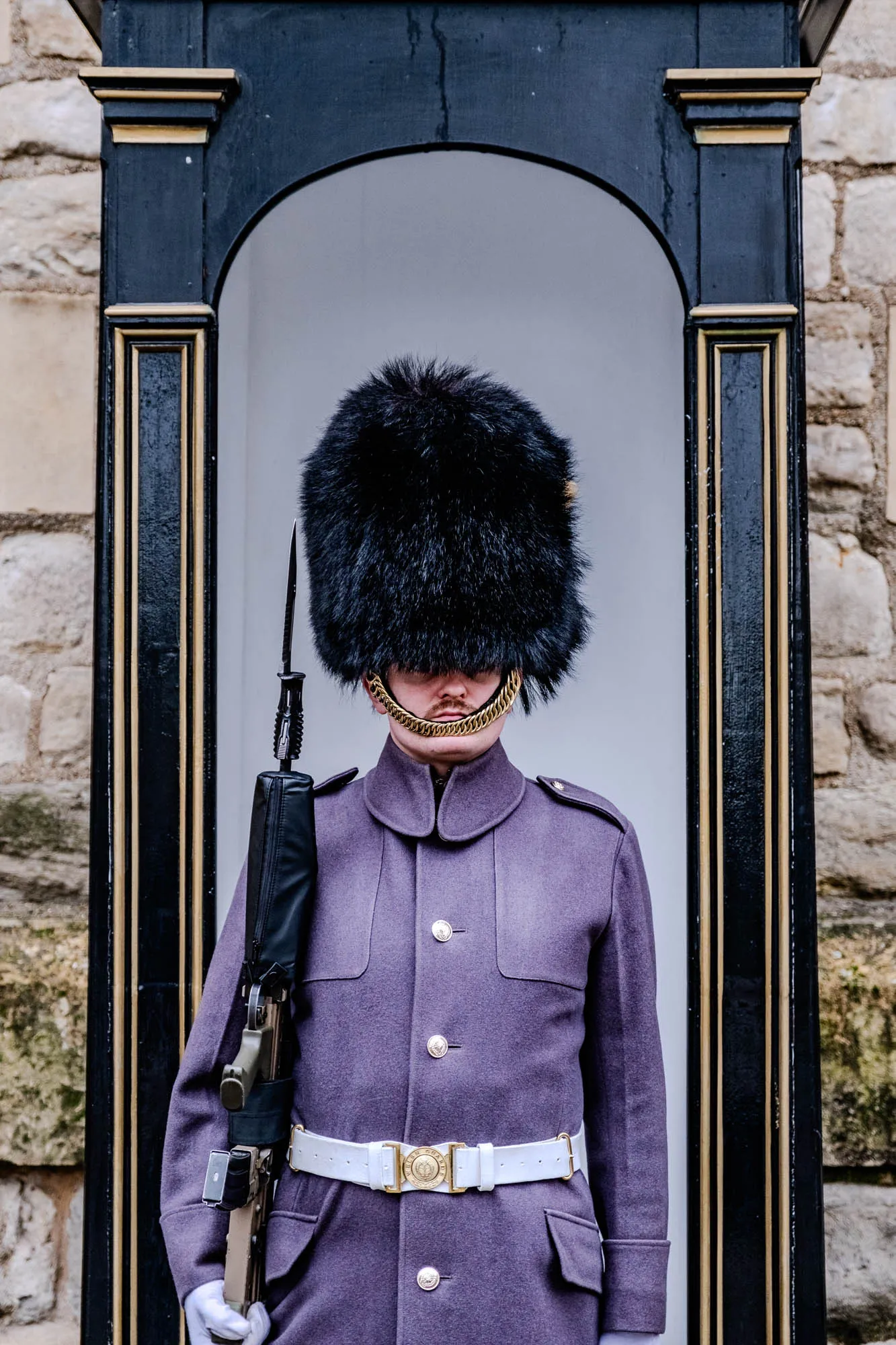 The image shows a person standing in a doorway. The person is wearing a dark purple jacket with gold buttons and a white belt. They are also wearing a black, furry hat that covers their eyes and a gold chain around their neck. Their right hand is holding a rifle that has a black strap and a long wooden barrel. The doorway has a black frame with gold trim, and the background is a stone wall.  