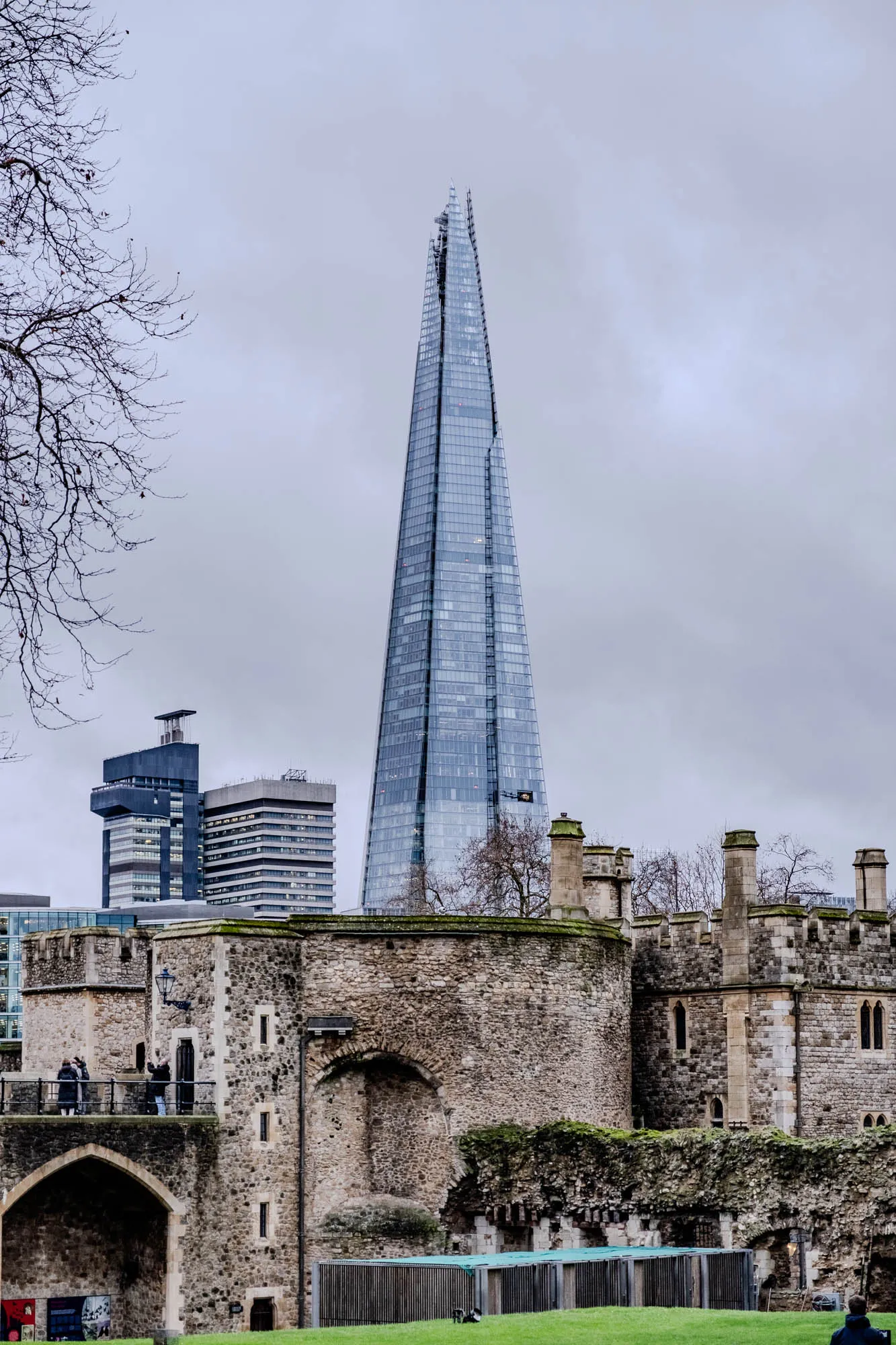 This image shows a tall modern glass building in the distance, with a sharp point, in front of a stone wall of an older building. The modern building is called the Shard and it is a skyscraper in London. The stone wall is part of the Tower of London, a historic castle and former prison. There are trees in front of the wall, and a few people standing on a walkway in front of the wall. The sky is gray and overcast.  A smaller office building is also visible behind the stone wall, and to the left of the Shard.  There is a grassy area in the foreground. 
