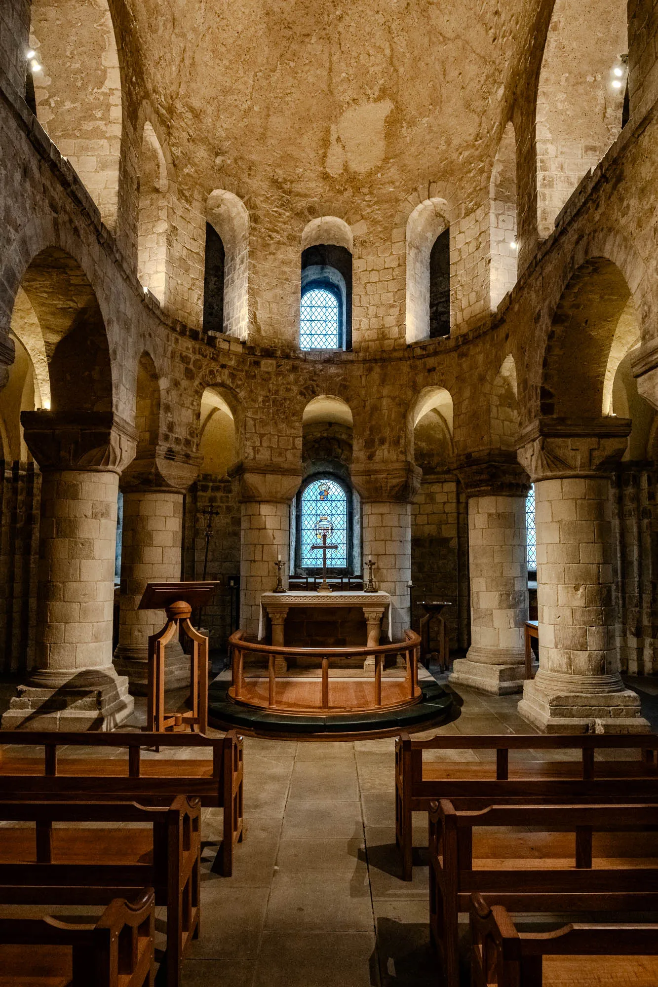 The image depicts a circular room with a domed ceiling. The room is constructed from stone, with numerous large arches and pillars. There are stained glass windows in three of the arches, and the room is lit with a dim, warm light. In the center of the room is an altar with two candles on it, with a cross above it. The altar sits on a raised platform, and there are wooden pews on either side of the altar. The pews are in the front of the room, closest to the viewer.  There are two more wooden pews in the room, one on the right and one on the left, away from the altar.  The floor is made of stone tiles. The room is a place of worship.