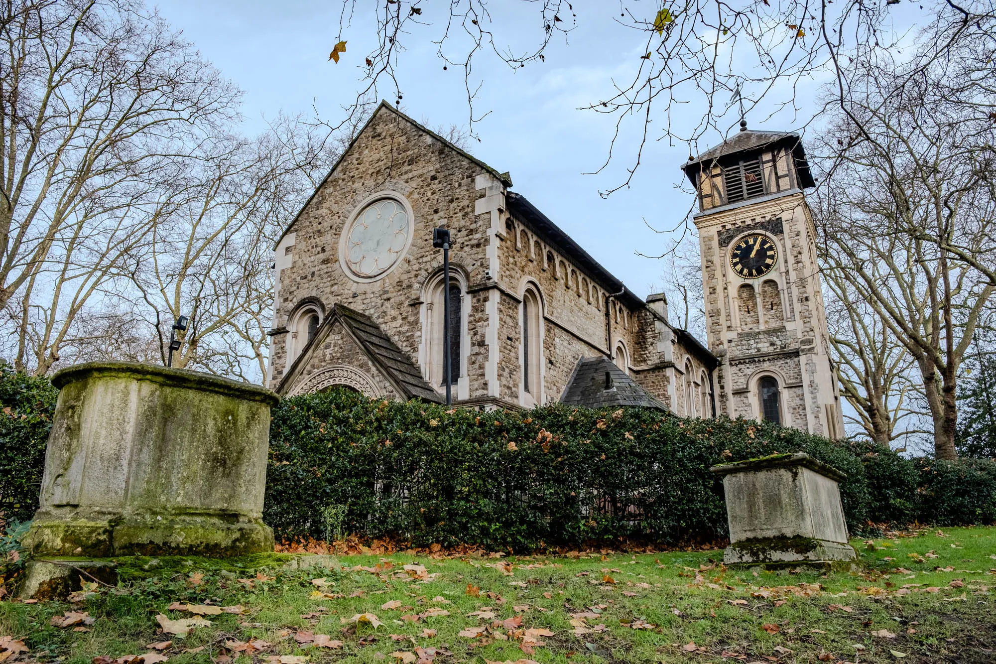 A low-angle view of a stone church with a tall clock tower. The church is built from rough-hewn stone with a slate roof. The tower is square and has a clock face on one side. The church is surrounded by a hedge and trees. The ground in the foreground is covered in fallen leaves. There are two stone monuments in the foreground, which are possibly tombs. The sky is a light blue with some clouds. The image is taken in autumn.