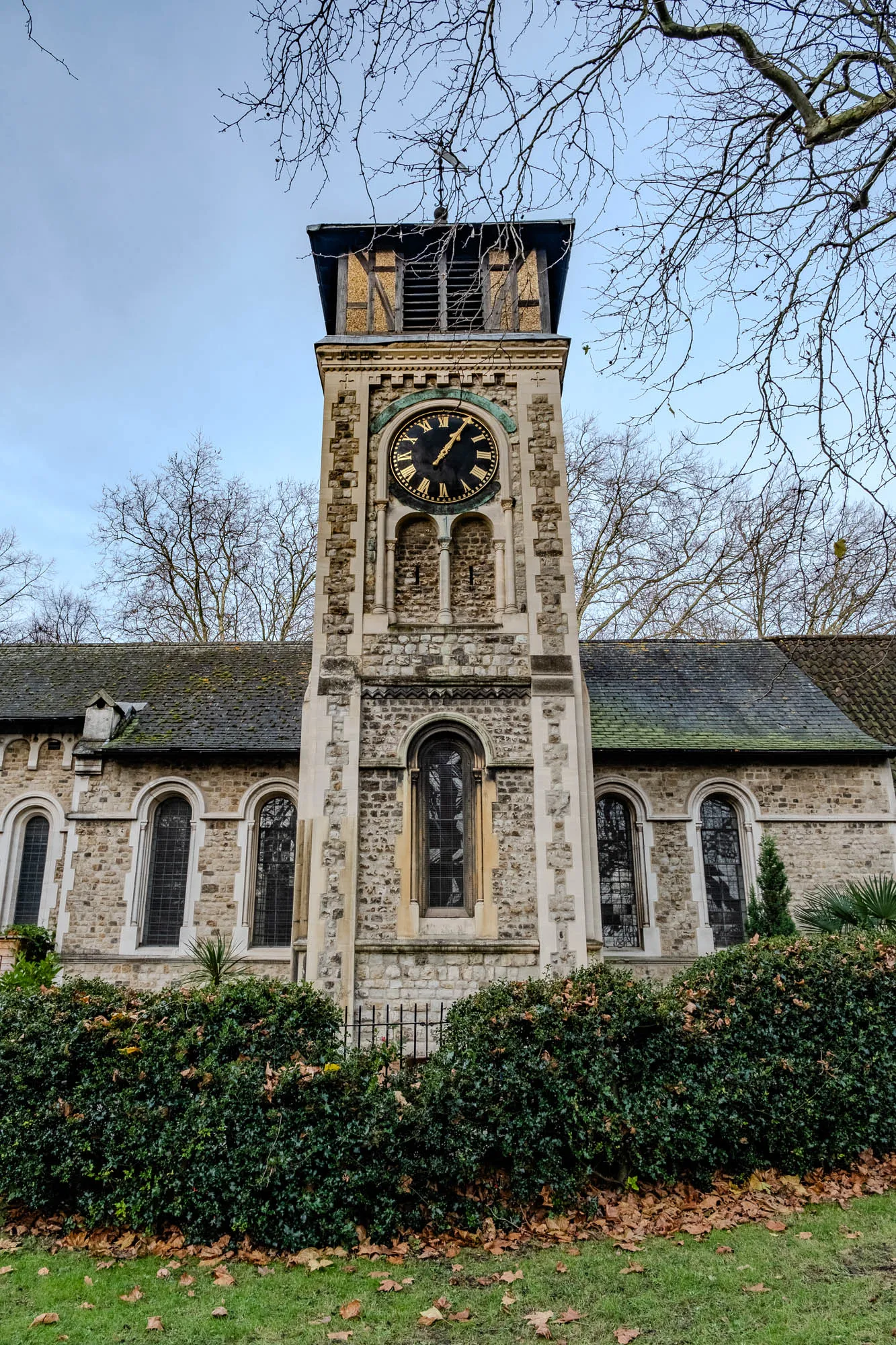 The image shows a stone church with a clock tower. The tower is tall and slender and is made of rough stone. It has a clock face with Roman numerals and gold hands. The clock is set against a pale blue sky. The church is partially obscured by a large bush of green leaves. The ground is covered in brown leaves and grass. The branches of a bare-branched tree are silhouetted against the sky. The church is a traditional building, with a slate roof and stone walls. There are large, arched windows on the sides of the church and small windows in the tower. The church is lit by the sun.