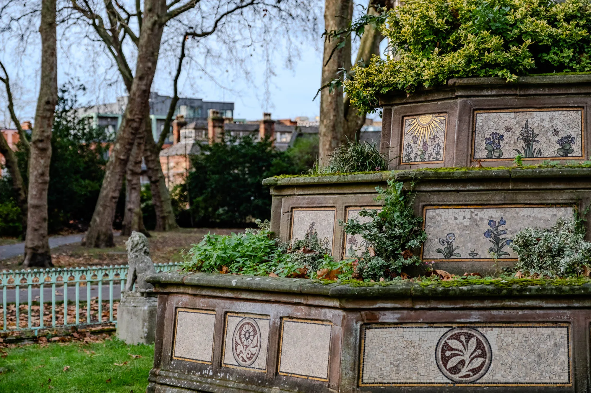 The image shows a stone structure with a mosaic design, likely a planter or a fountain. It's located in a park or garden setting, with lush green grass in the foreground and several bare-branched trees in the background. The structure is made of several layers, and each layer has a mosaic of colorful flowers and leaves. The top layer features a mosaic of a sun with rays of light.  The bottom layer features a mosaic of a plant with leaves and a circle surrounding it. There is a stone lion statue on the left side of the image, which is partially obscured by a green metal fence.  The structure is covered in moss and is weathered with time.  The background is blurred and shows a cityscape with buildings in the distance.