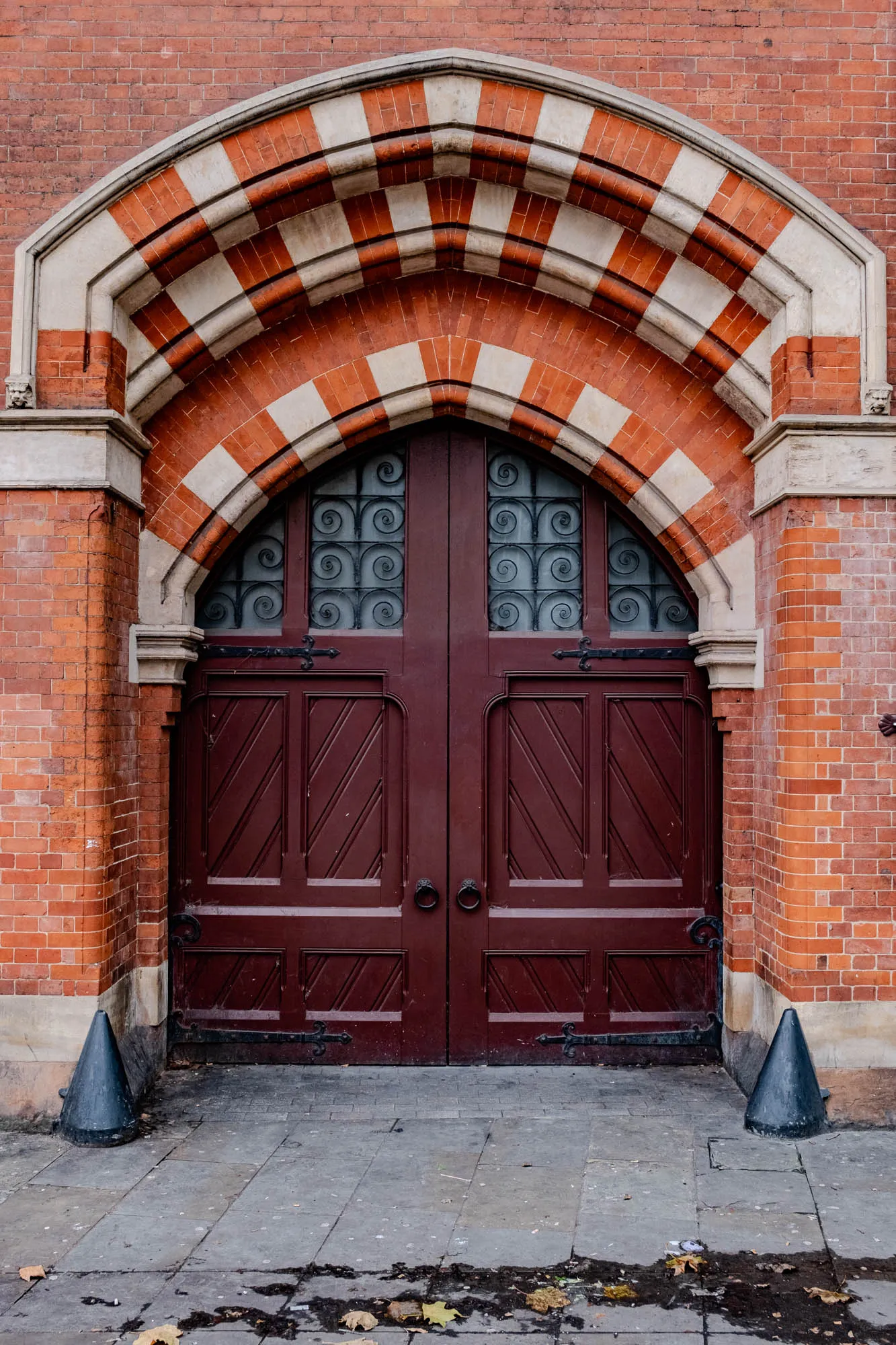 A brick building with a large arched doorway. The arch is made of red bricks and white mortar, arranged in a repeating pattern of red, white, red, white, etc.  The archway is framed by a pair of double doors made of dark wood. The doors have a  decorative iron design on the top panels and diagonal wood panels below. There are two large, black iron hinges on each door. The doors have large, round, black iron handles with ring pulls in the center. There is a gray sidewalk in front of the doors. There are two black, cone-shaped objects lying flat on the sidewalk to the left and right of the doorway.  The sidewalk is uneven and has patches of dirt and leaves on it.