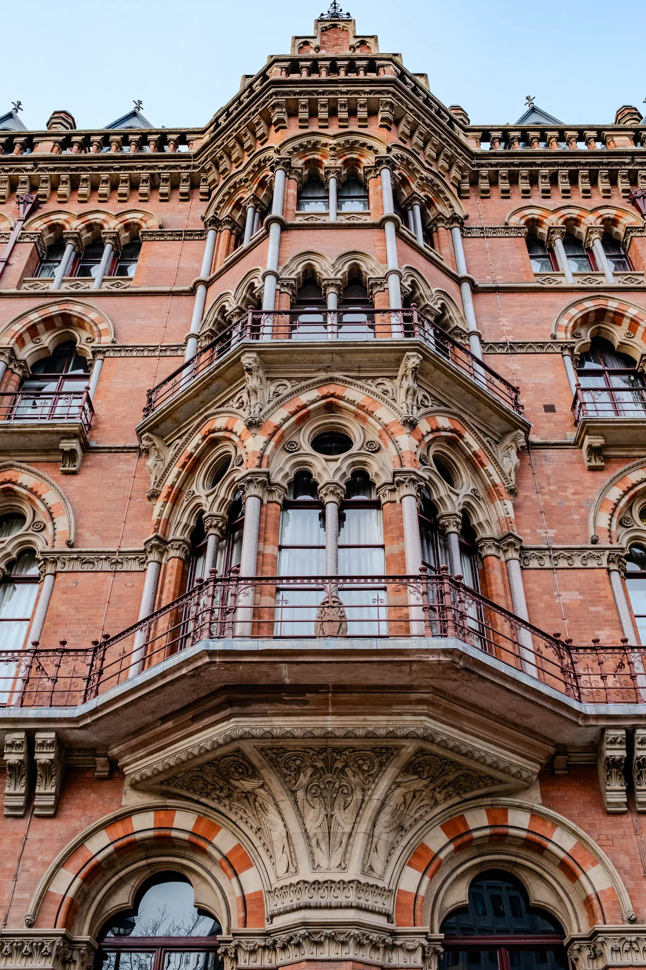 This image is a low angle shot of the exterior of an ornate building with red brick and stone. The building is tall and the image only shows the top few floors. The building features many arched windows, some of which are topped with decorative stonework. The windows have multiple panes of glass and there is an intricate wrought iron balcony around each of the windows. The windows feature ornate stone trim and look to be about 3 stories high.  The building has a large decorative cornice that runs the entire length of the building, and it has a distinctive cornice with a large and intricately carved stone decoration at the bottom.  There is also a decorative stonework arch with an ornate stonework design. The building is brightly lit and the sky in the background is a clear blue.  The building looks to be very old and well-maintained.