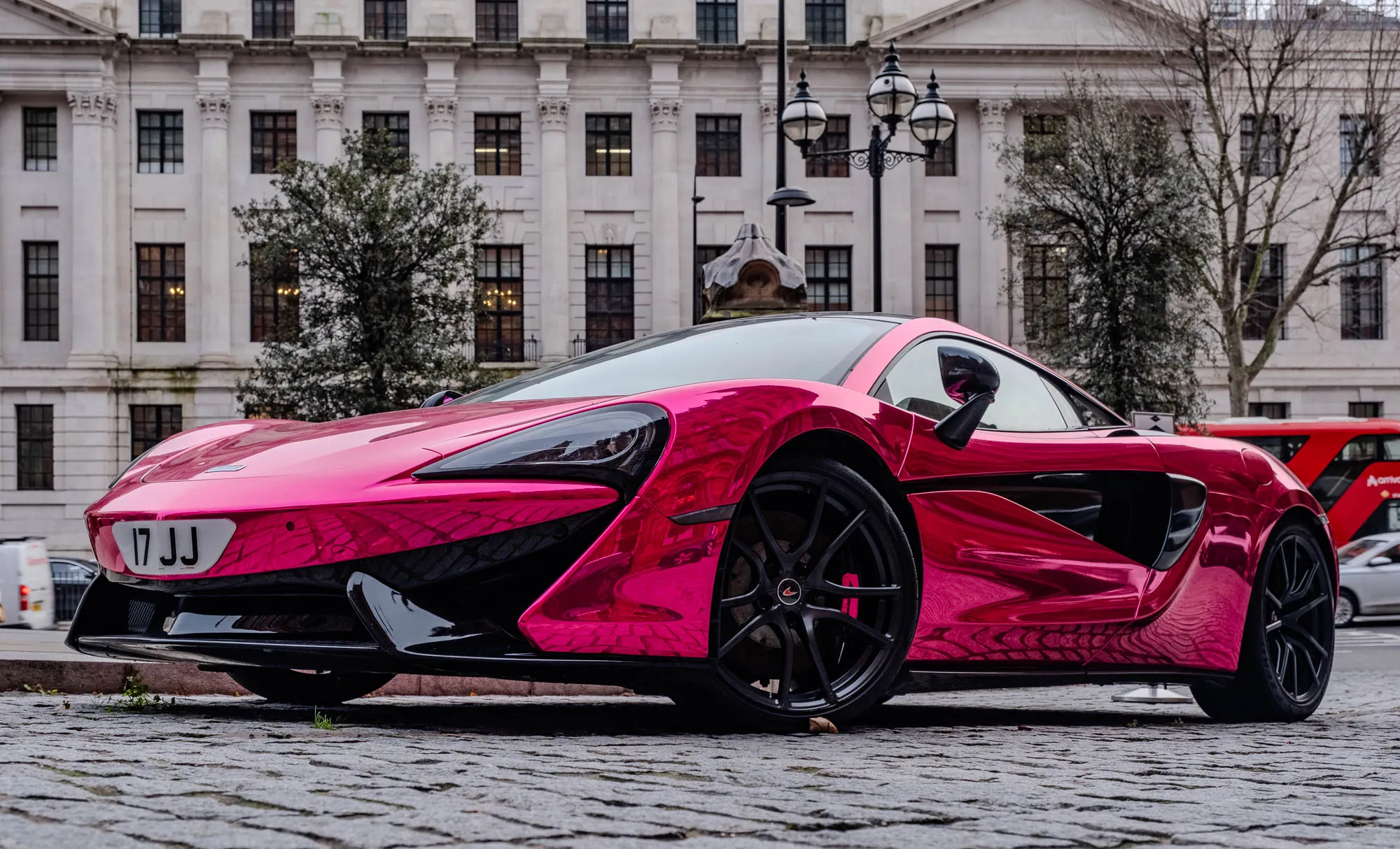 The image shows a bright pink McLaren 570S parked on a cobblestone street in front of a tall white building with many windows. The car has a black front bumper, black side skirts, and black wheels with red brake calipers. The car has a black side mirror. The license plate reads "17JJ".  A street light with black and gold accents is partially visible in the background. In the background, on the right side of the image, a red double-decker bus is partially visible and a white car is parked further down the street. The building has a white stone facade. It has many windows and a decorative stone element on the roof. The image is taken from a low angle, looking up at the car. The image is well-lit, and the colors are vibrant.