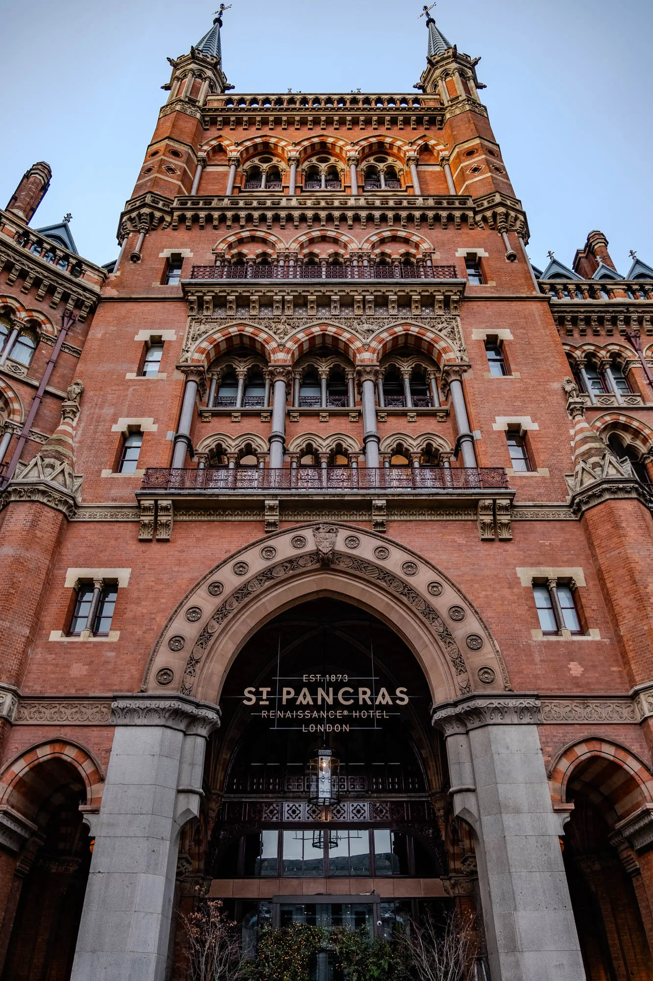 The image is of a tall, red brick building with ornate detailing. The building has multiple arched windows and a large arched entranceway in the center. The entranceway is framed by two large pillars and above it is the sign "Est. 1873 St. Pancras Renaissance Hotel London." The building is highly detailed with many intricate carvings. The building is located in London, England. It appears to be a historical building that has been well-preserved.