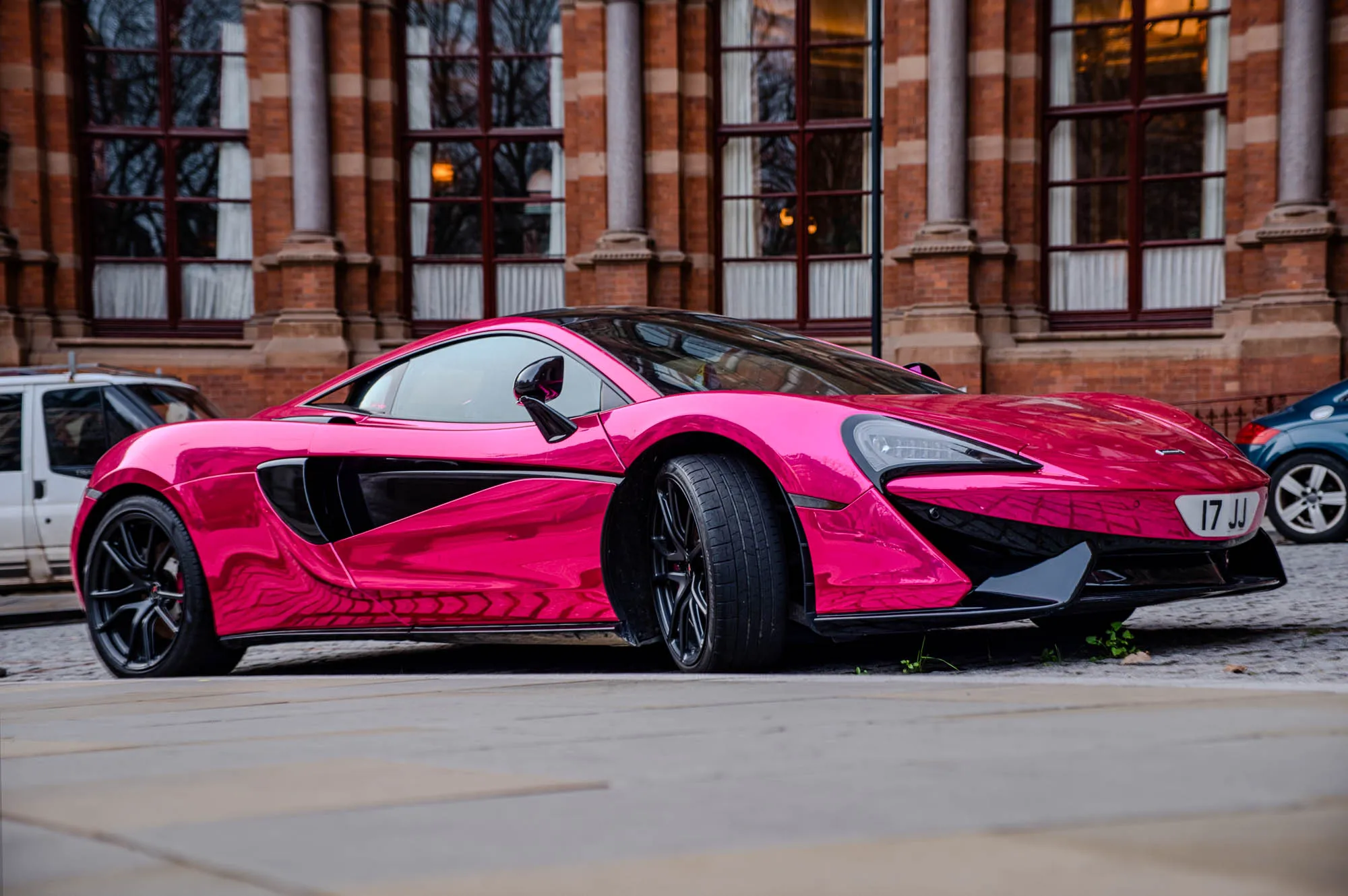 The image shows a bright pink McLaren 570S parked on a sidewalk in front of a brick building. The car is facing towards the right of the image with its front left wheel and part of the front bumper visible. The car has black accents on the doors and the back of the car. The license plate on the back is visible, and it says 17 JJ. The car is shiny and sleek, and the pink color is very vibrant. There is a white van partially visible to the left of the image. The brick building behind the car has large windows with white curtains and a tall column in front of the windows.