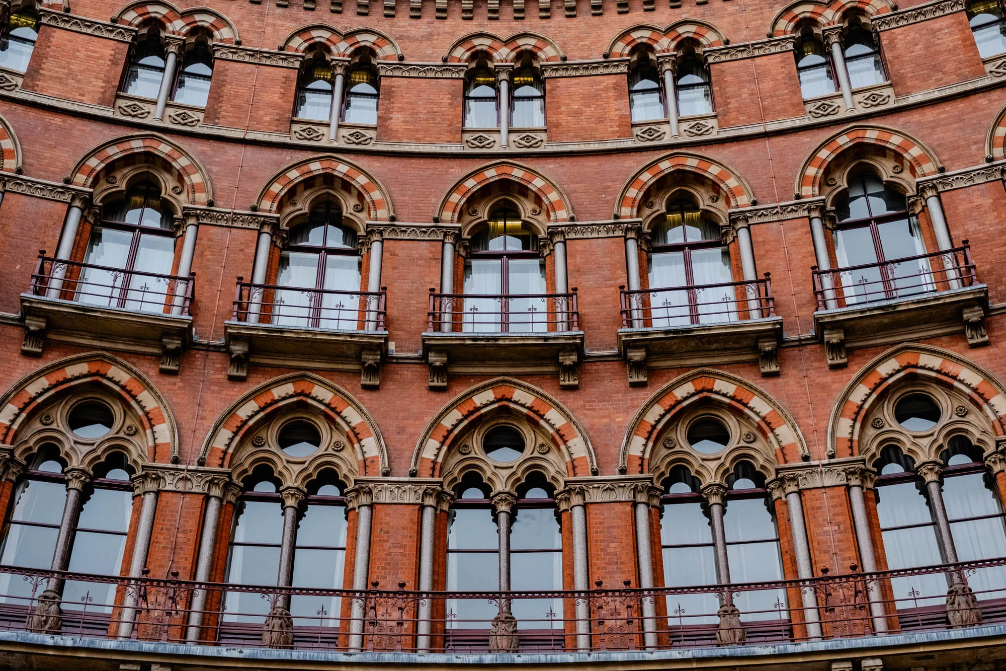 This image shows a portion of a red brick building with multiple arched windows. There are four levels of windows, all framed with intricate stonework. Each window has a small balcony with a metal railing. The windows are all rectangular and have white panes of glass. The building has a slight curve, making the bottom level of windows appear wider. The photo was taken from a low angle, giving the building a dramatic perspective. 
