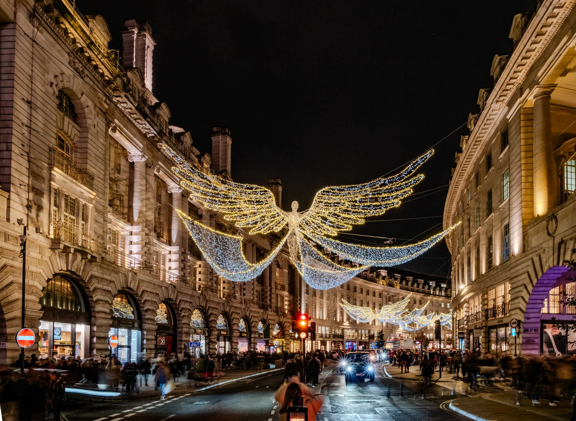 The image depicts a nighttime street scene with a large, illuminated angel sculpture strung with lights across the street. The angel is made of a white frame with small white lights outlining the shape and golden lights filling in the wings and body. Below the angel, strings of white lights with some gold interspersed are strung between buildings on both sides of the street, creating a canopy of lights. The buildings on both sides of the street are tall, ornate, and made of stone. They are lit from within and have large windows, some of which are displaying products for sale. The street is crowded with people, many of whom are blurred in the image, walking and shopping. The image has a festive and celebratory feel.