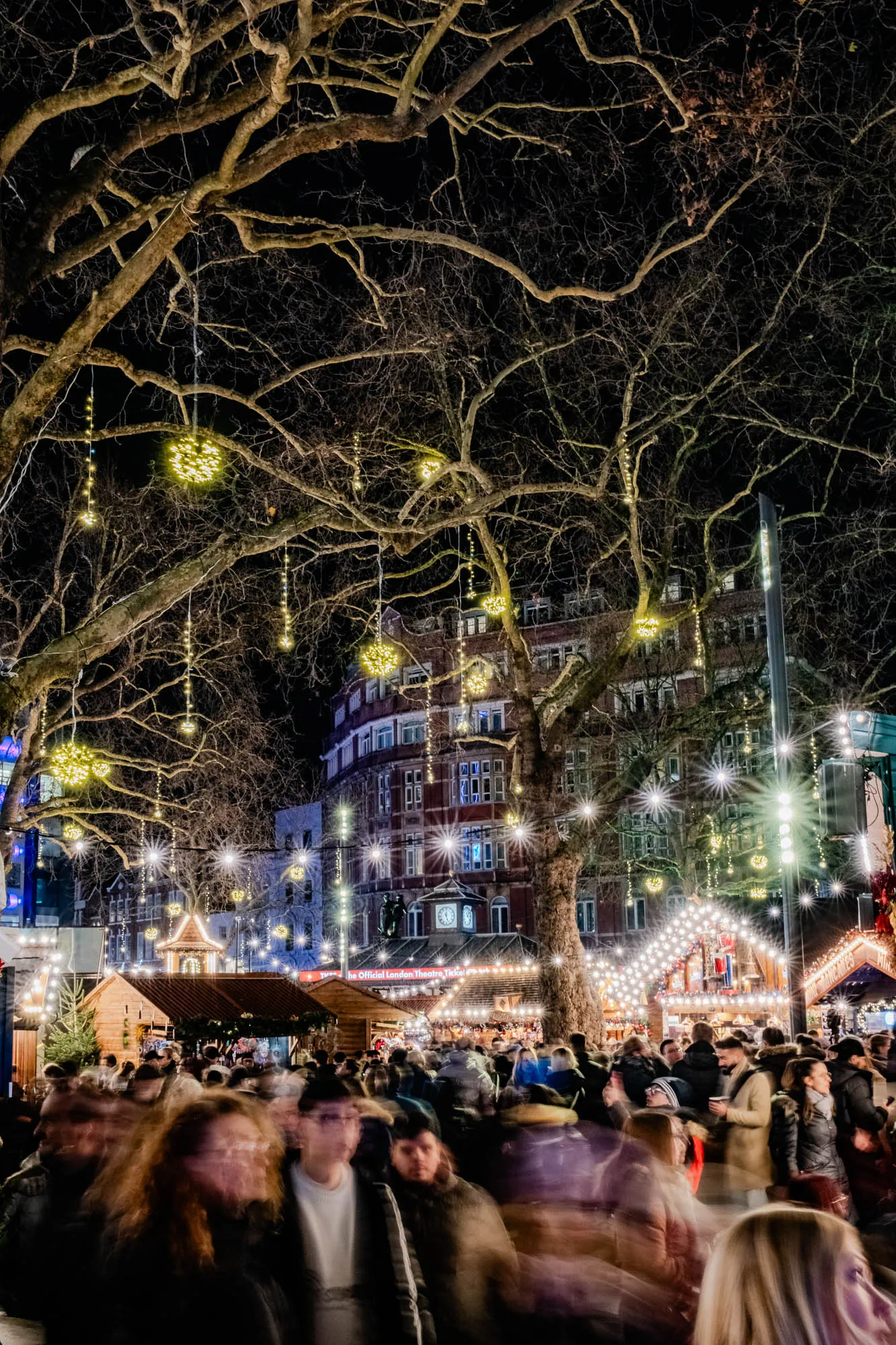 The image shows a night scene with a large tree in the center, its branches laden with decorative lights in the shape of balls. The tree is silhouetted against a dark sky, and the background is blurred but appears to be a cityscape. The tree is adorned with strings of small white lights that cascade down from its branches. The tree is in the foreground, and behind it, a large building can be seen. The building is lit up with warm yellow light, which creates a cozy and inviting atmosphere. In front of the tree, many people are walking in a busy street. The people are blurred and moving quickly. This indicates a bustling, lively scene, possibly a Christmas market or a festive gathering. The image captures the joy and warmth of the season, with its bright lights and cheerful crowd.