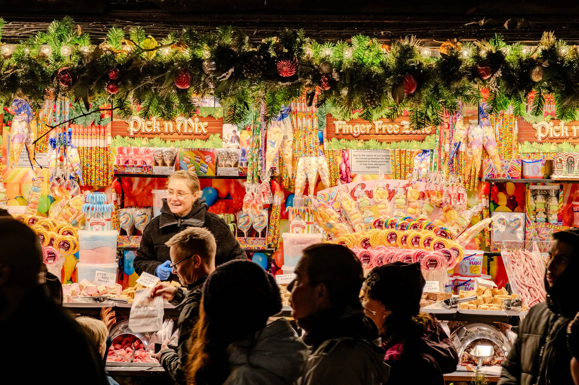 A woman selling candy from a well-lit winter market stall. The stall is filled top to bottom with candy. People walk past the stall and are in shadow. There are two signs visible: "Pick n Mix" and "Finger Free Zone".