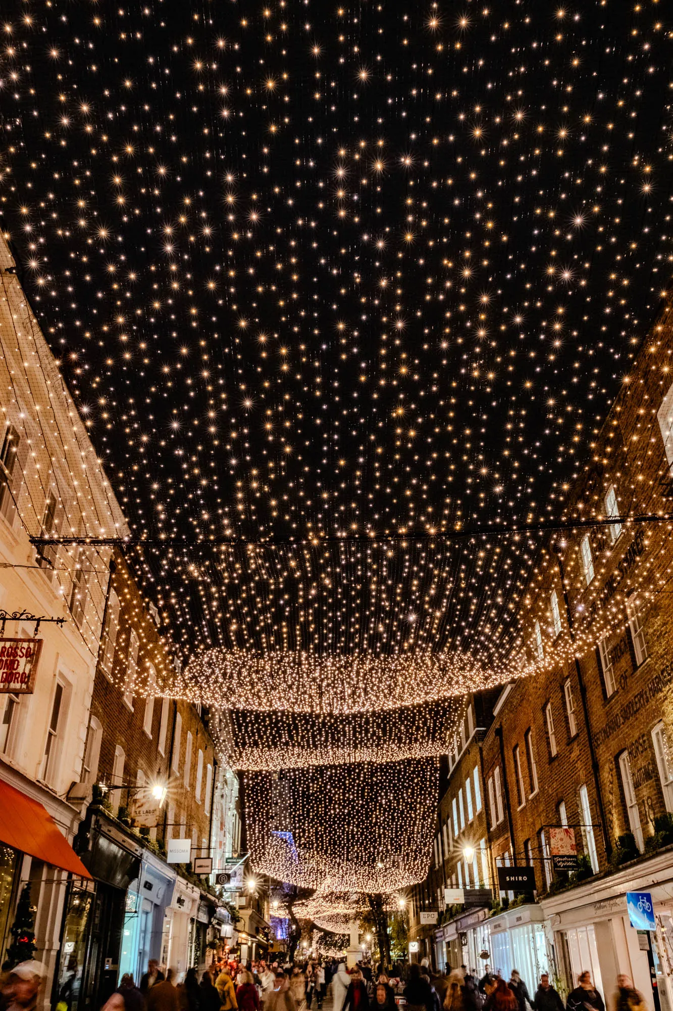 The image shows a street at night with a canopy of twinkling lights above. The lights are strung across the street and create a sparkling, starry effect. There are buildings on either side of the street, and the windows of the buildings are lit up. There are people walking in the street, but they are blurry and hard to make out. The scene is romantic and festive, and it evokes a sense of joy and wonder. 
