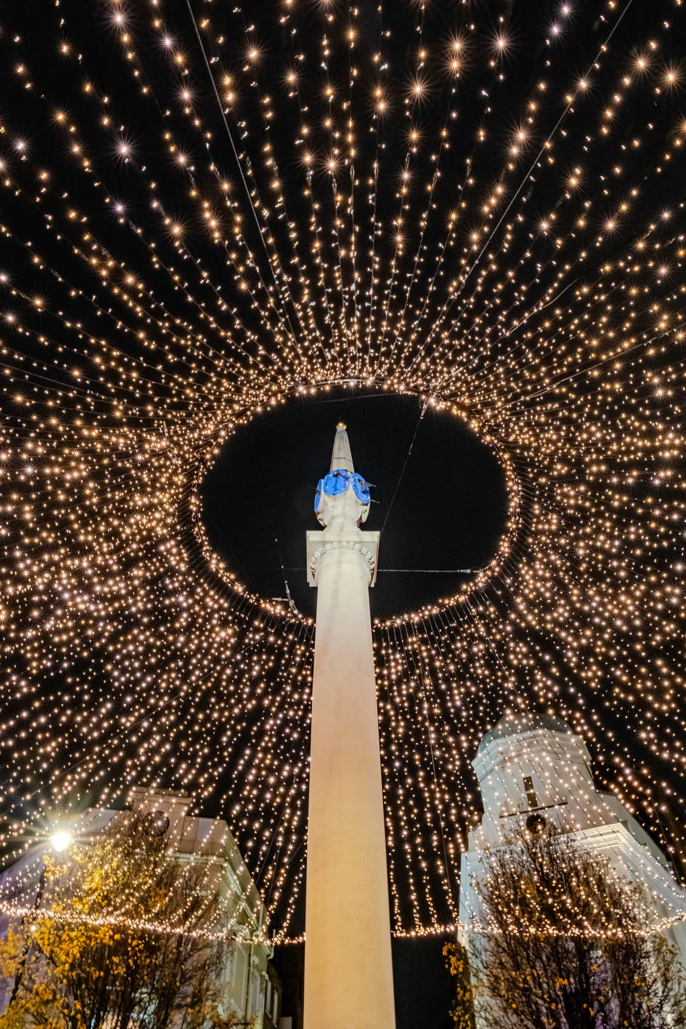 The image shows a tall white pillar with a blue cap on top, standing in the center of a circular arrangement of string lights. The lights are strung from building to building and arch over the top of the pillar. There are hundreds of tiny twinkling lights, creating a canopy of light against the dark night sky.  The lights are arranged in a symmetrical pattern, forming a star-like shape above the pillar.  In the background, two buildings are visible. One appears to be a church with a cupola on top. There is a tree on the right side of the image.  The scene is reminiscent of a festive and celebratory atmosphere. 
