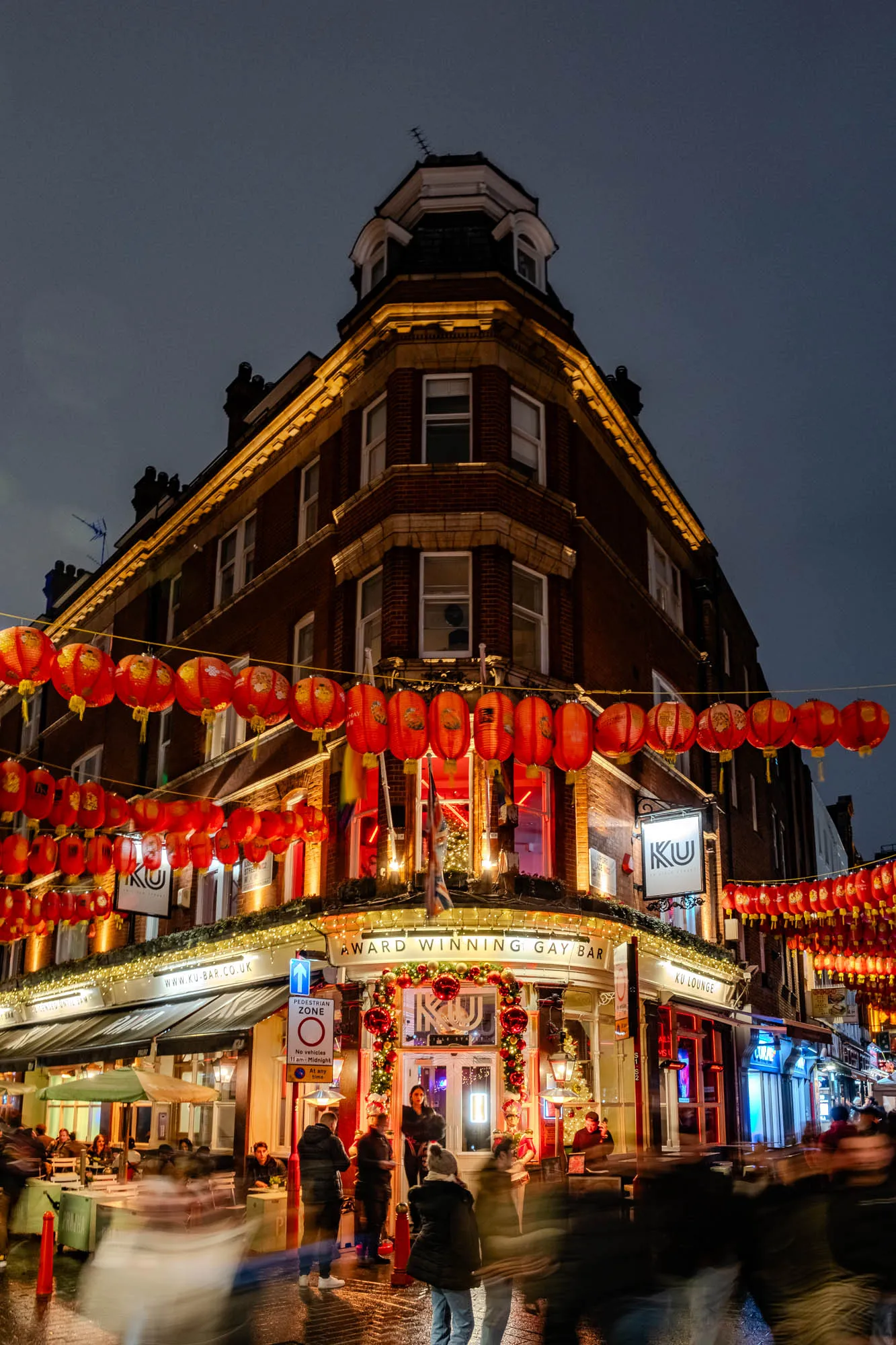 The image shows a brick building with a sign that says "KU Award Winning Gay Bar". The building is decorated with red Chinese lanterns, Christmas lights, and greenery. There is a sign that says "KU" above the entrance to the bar, and another sign that says "KU Lounge" above the entrance to a separate area of the bar. The street is lit up, and there are people walking around. In the foreground there is a sign that indicates pedestrian zone and is lit up by a light. There are blurry figures of people walking through the street.  The sky is dark and the building is brightly lit.