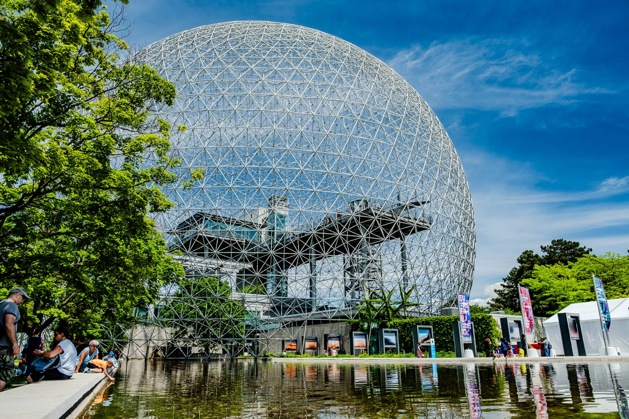 The image shows a large, metal geodesic dome against a bright blue sky with white clouds. The dome is made of a lattice of interconnected metal beams, creating a complex geometric pattern. The dome appears to be partially surrounded by trees. There are people standing on a platform high above the ground near the top of the dome. In the foreground, several people are standing and sitting on the edge of a reflecting pool, which is mirroring the dome and the sky above.  To the right of the dome there are some white tents and colorful signage.  A leafy green tree hangs over the left side of the image.