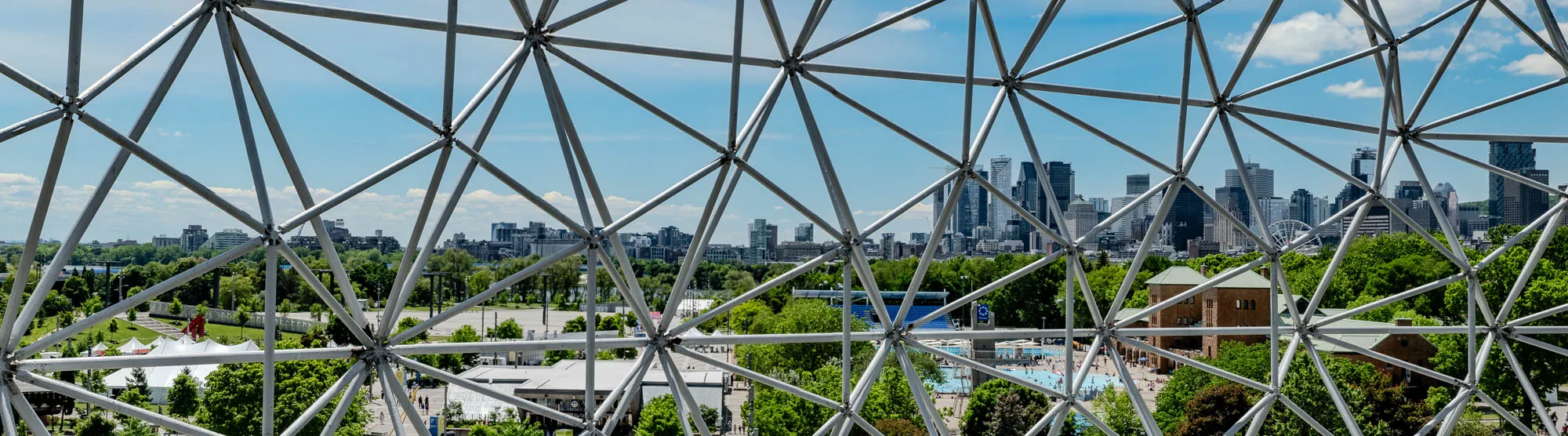 The image shows a view through a large, geodesic dome-like structure. The frame of the structure is made of thin, silver metal bars that form triangles. Through the frame, you can see a city skyline in the distance. The city consists of several tall buildings, primarily skyscrapers with some smaller buildings scattered around them. In the foreground, there are trees and some green, grassy areas, along with a few white tents. The sky is a clear, bright blue with a few puffy white clouds.  To the right of the cityscape, there is a building with a brown brick facade and a green roof. It appears to be a public building or community center. The image conveys a sense of modern architecture and urban life, with the geodesic frame adding a unique and futuristic touch.